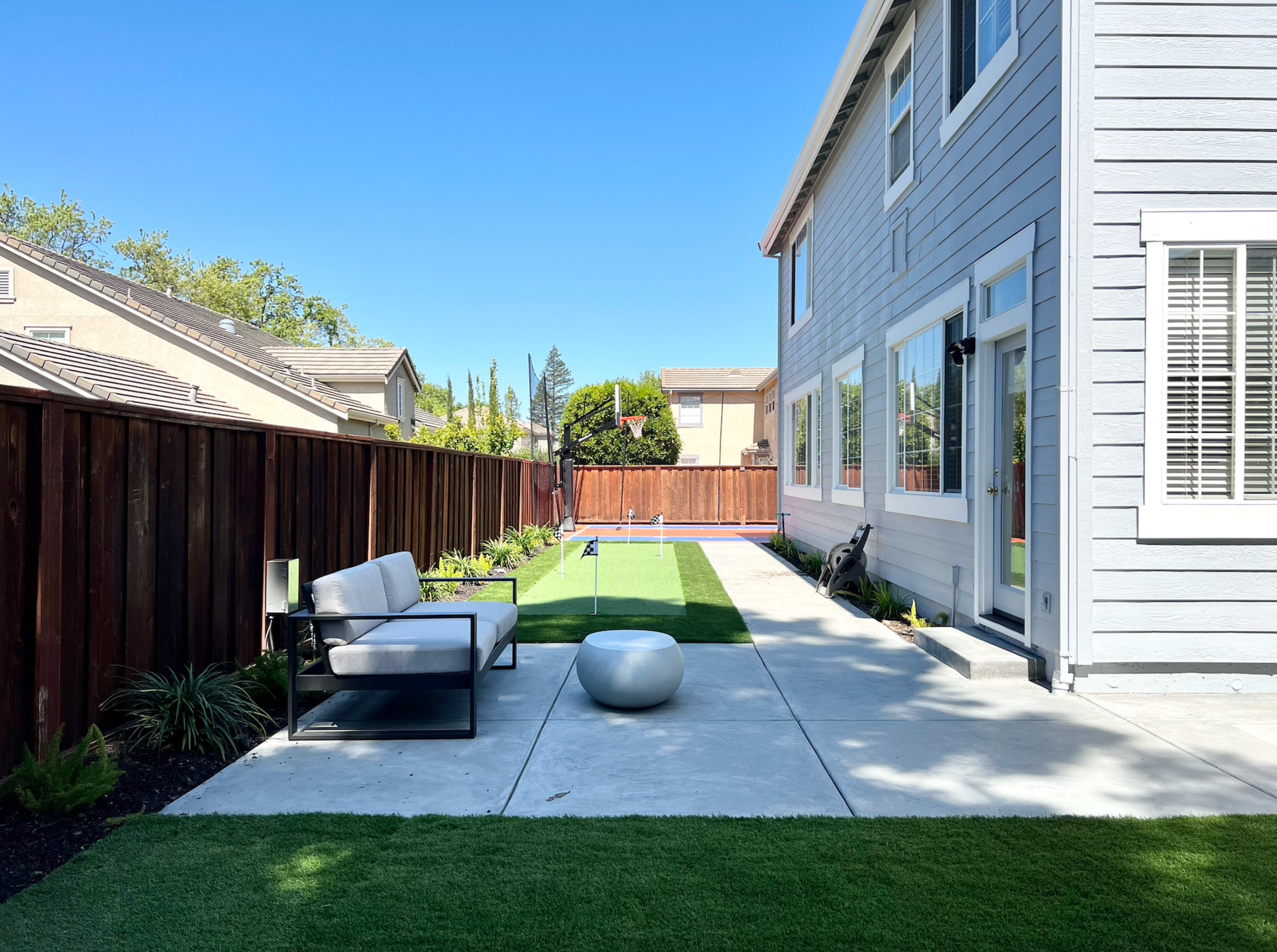 A well-maintained backyard with a concrete patio, green grass, and a modern seating area beside a wooden fence.