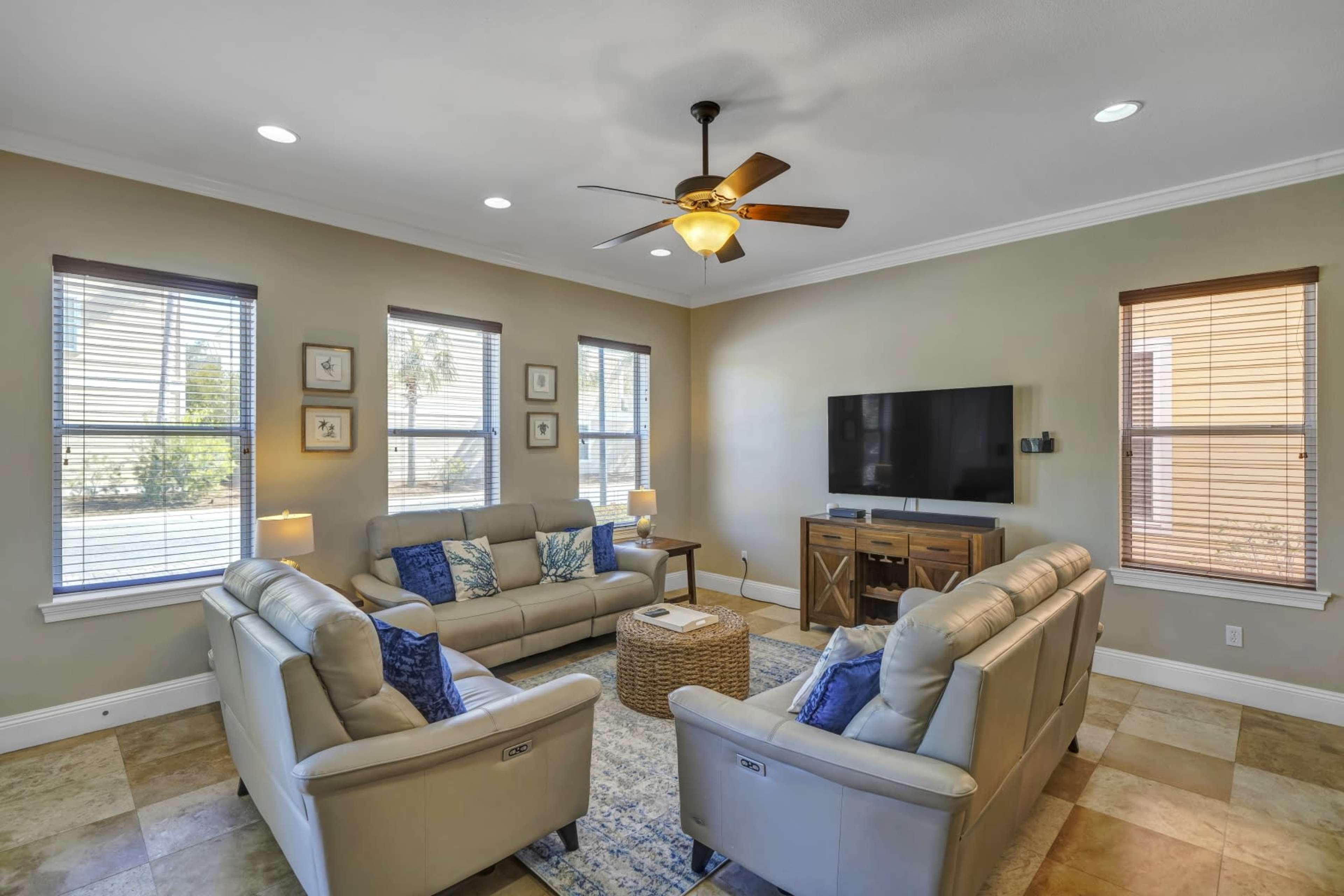 The living room features two couches facing a television, with a coffee table and a woven rug in the center, illuminated by natural light from the windows.