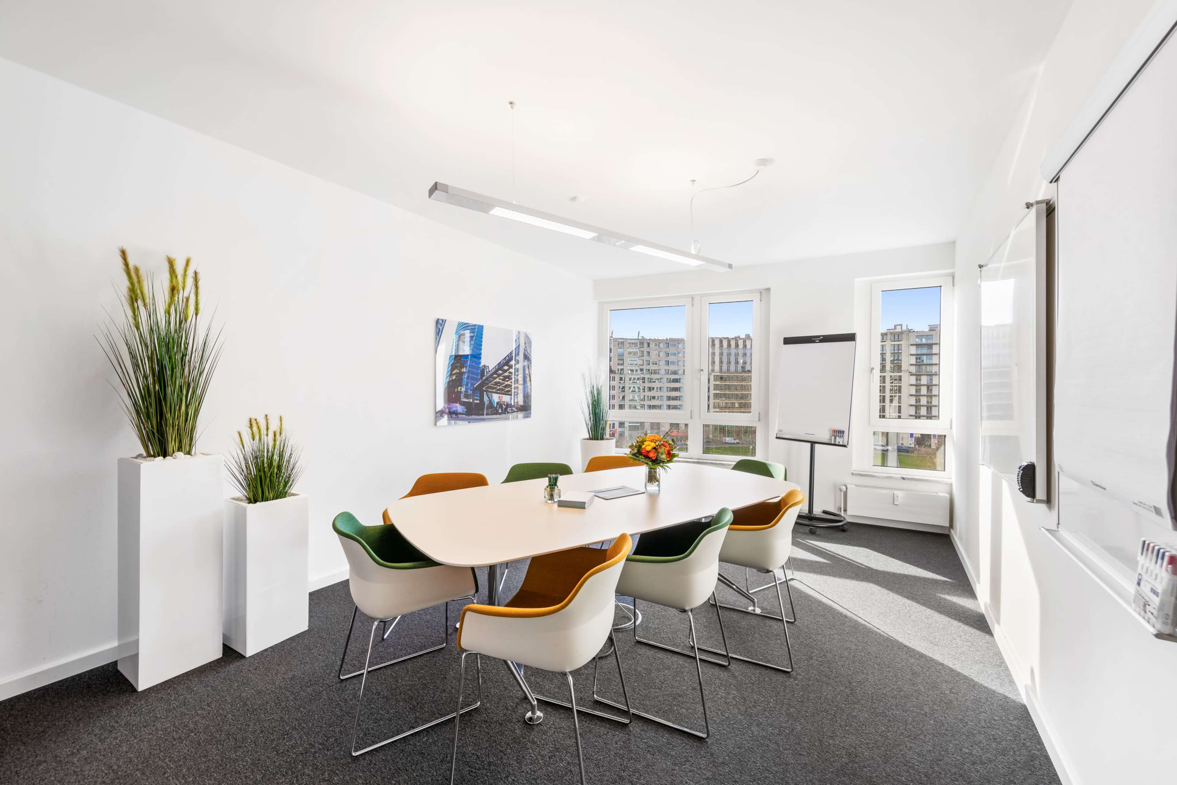 A modern conference room features a round table surrounded by six chairs, with large windows providing natural light and views of a cityscape.