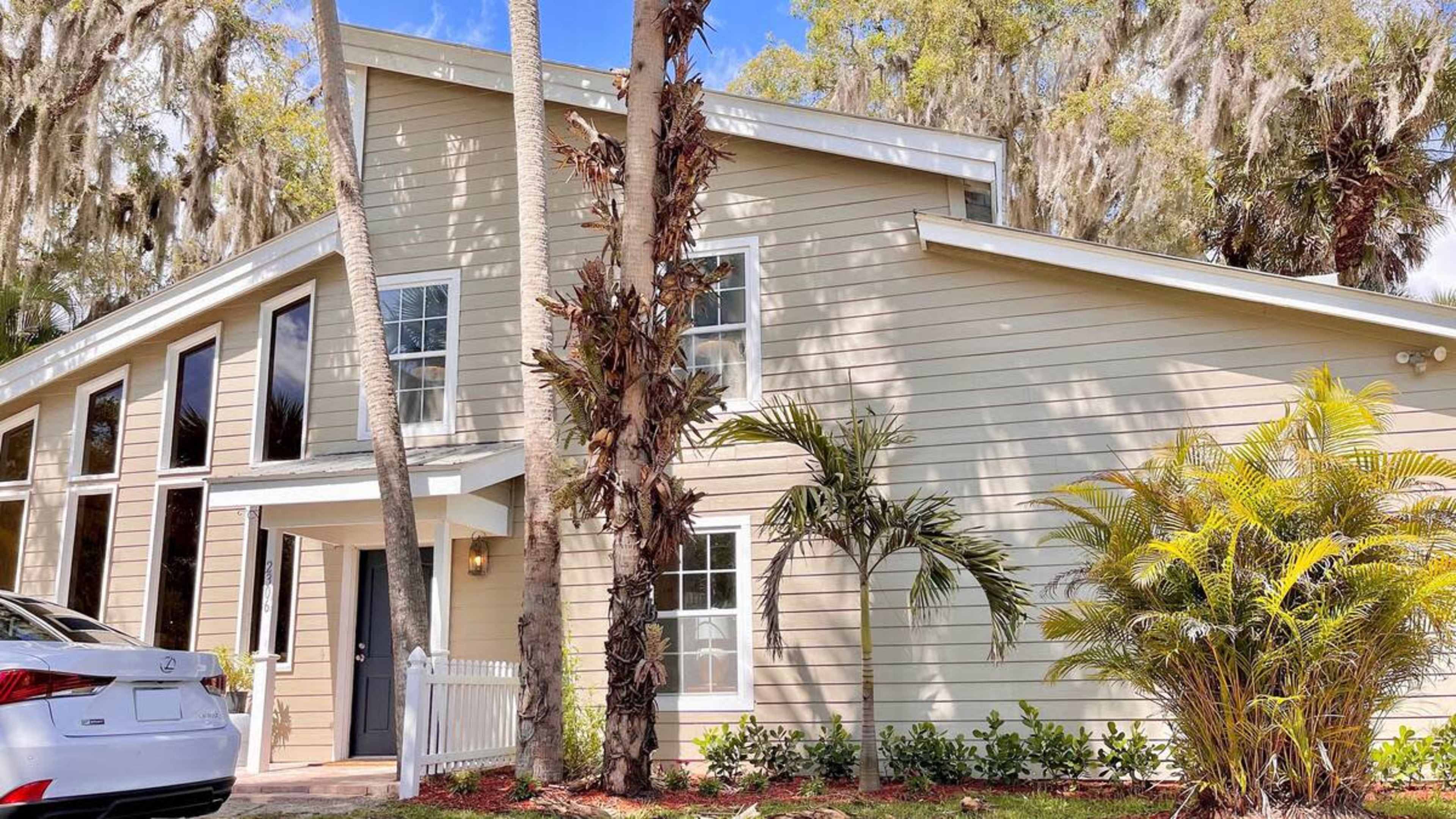 A two-story house with large windows and a front porch is surrounded by palm trees and shrubs.