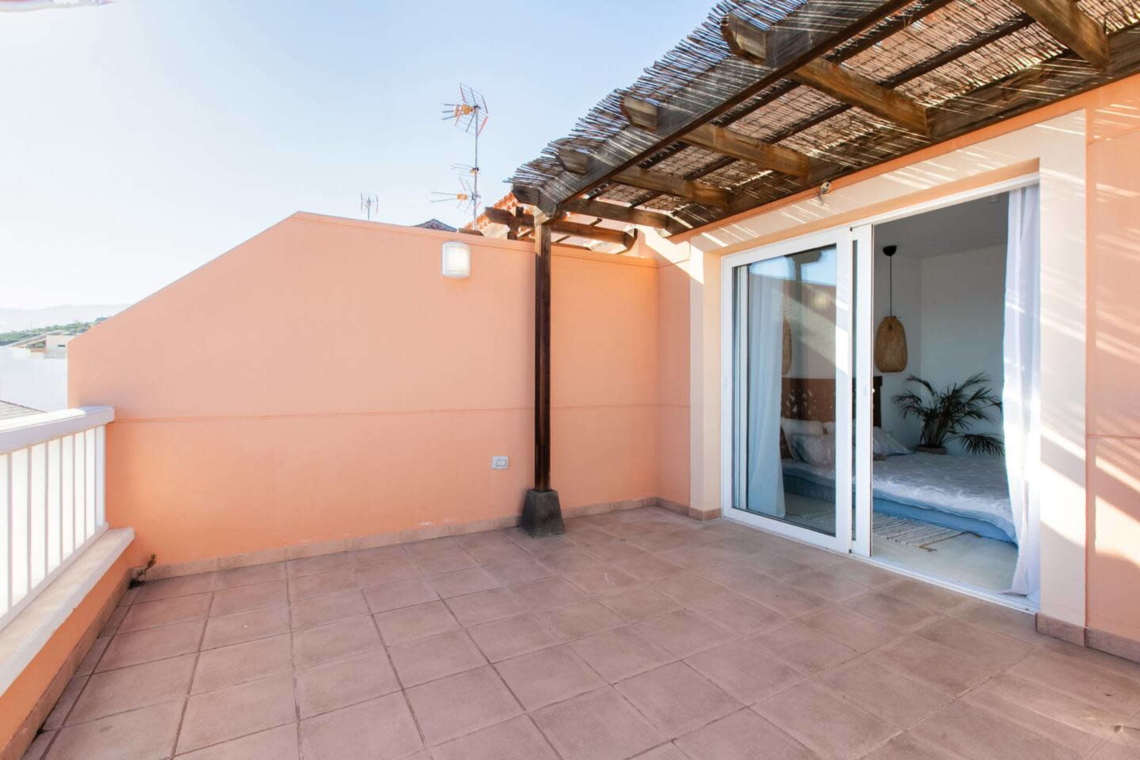 The image shows a spacious terrace with a tiled floor, a wooden pergola overhead, and sliding glass doors that lead into an interior room.