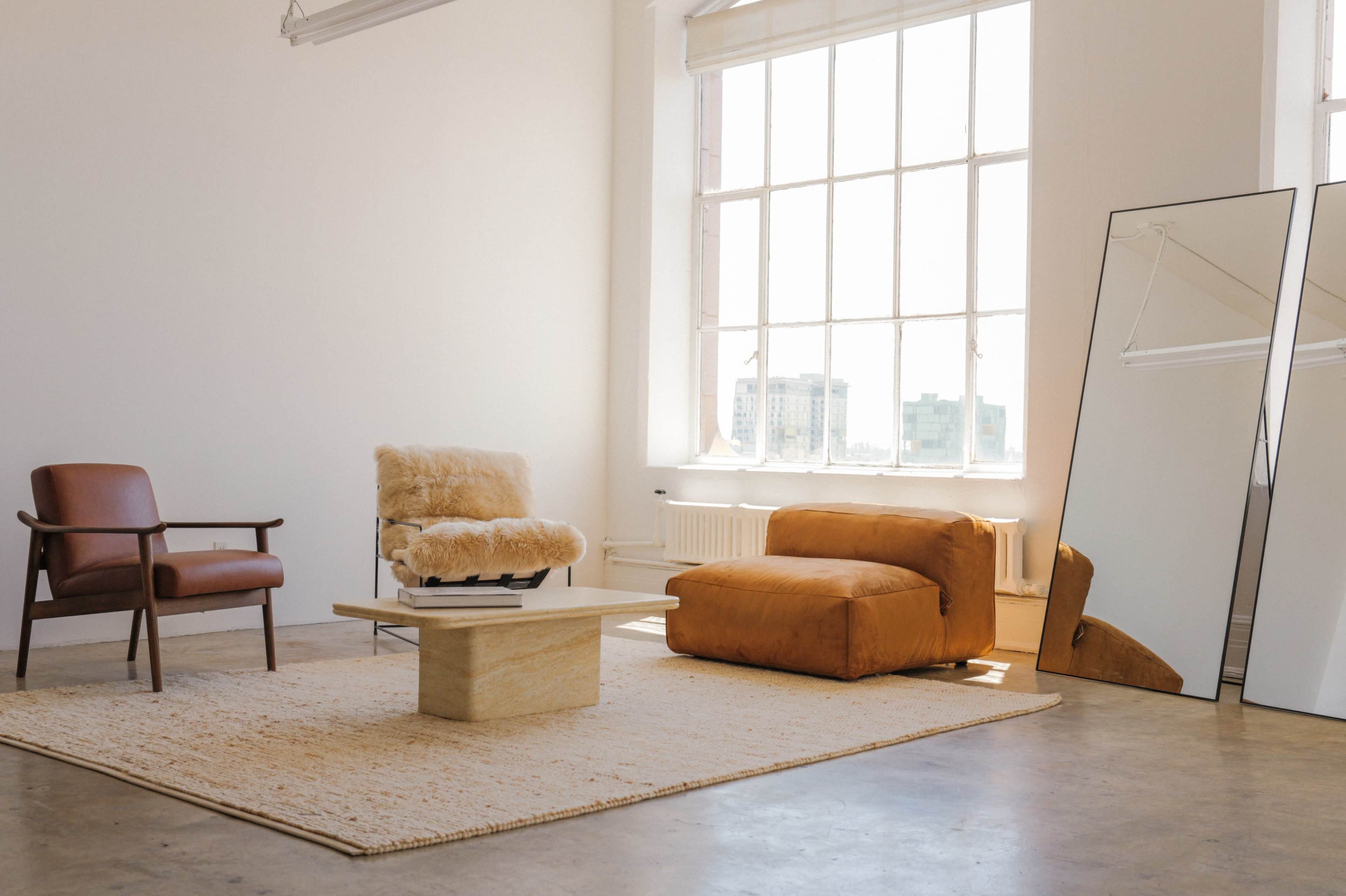 A modern, minimalist living room features a plush chair and a large orange lounge chair beside a wooden coffee table on a textured rug, with large windows allowing natural light to fill the space.