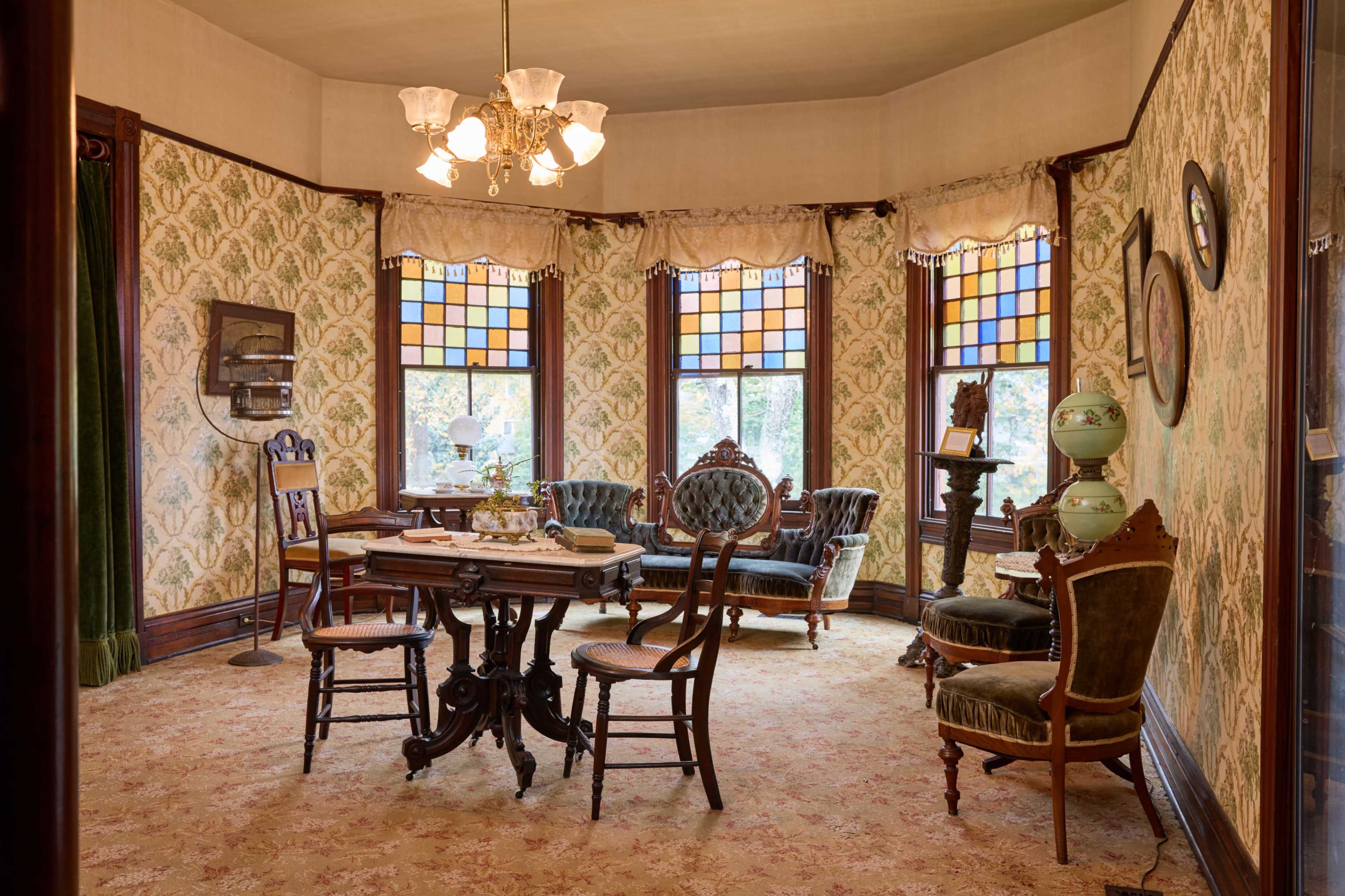 The image shows a vintage dining room with patterned wallpaper, stained glass windows, and antique furniture including a round table and upholstered chairs.