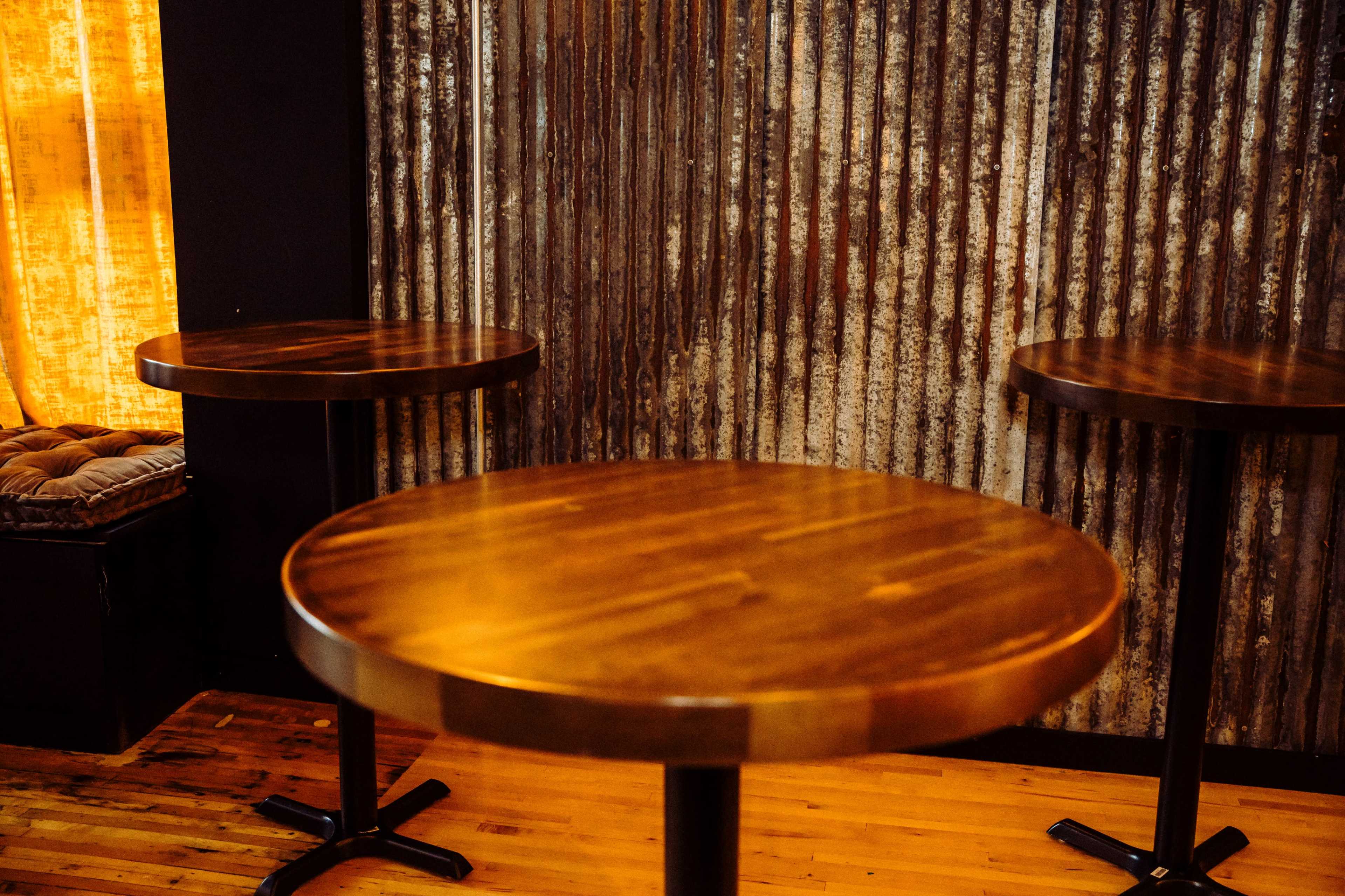 The image shows three wooden tables with metal bases arranged in a dimly lit space featuring a corrugated metal wall in the background.