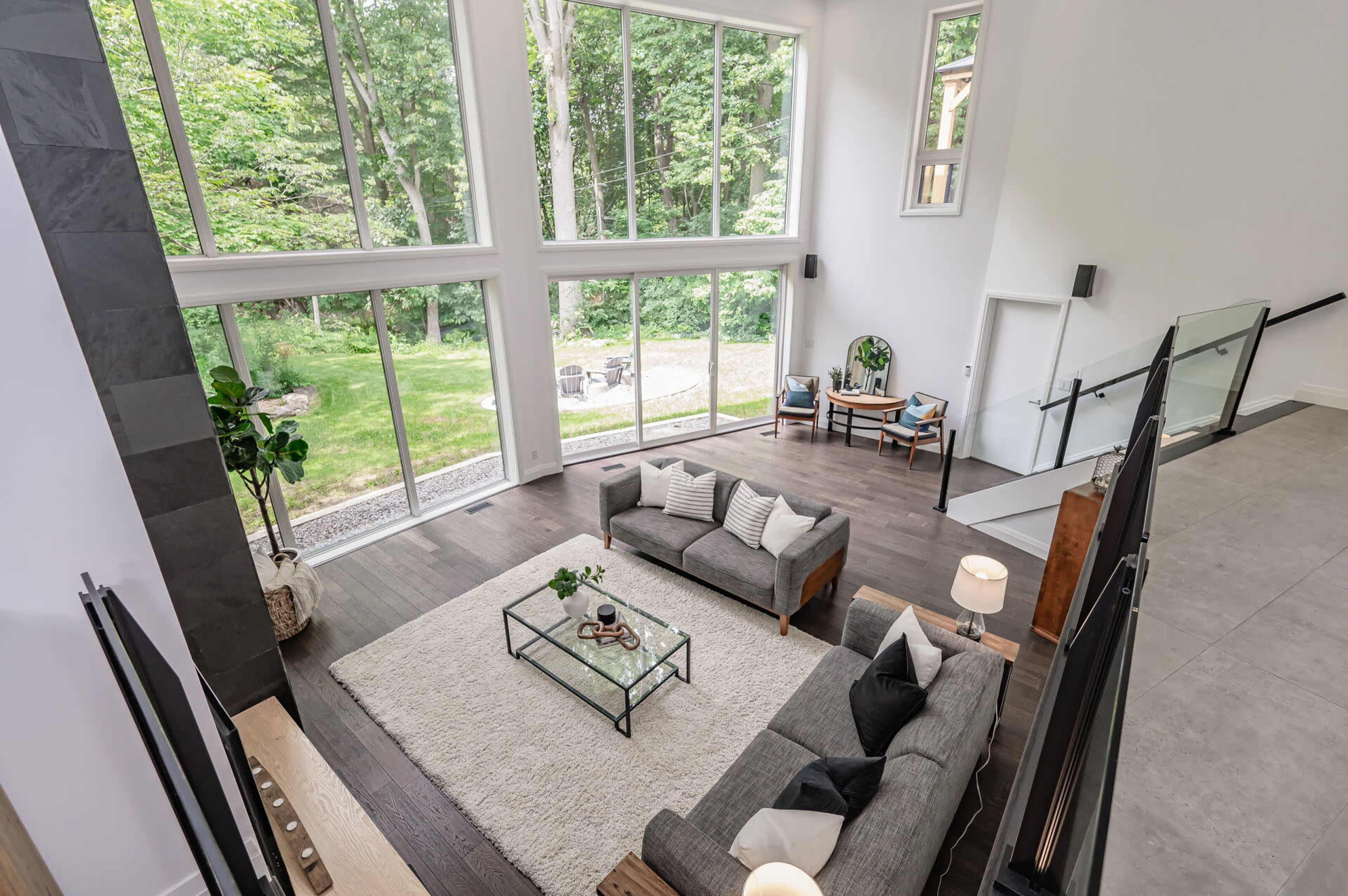 A modern living room with large windows overlooking a wooded area, featuring a gray sectional sofa, a coffee table, and a light rug.