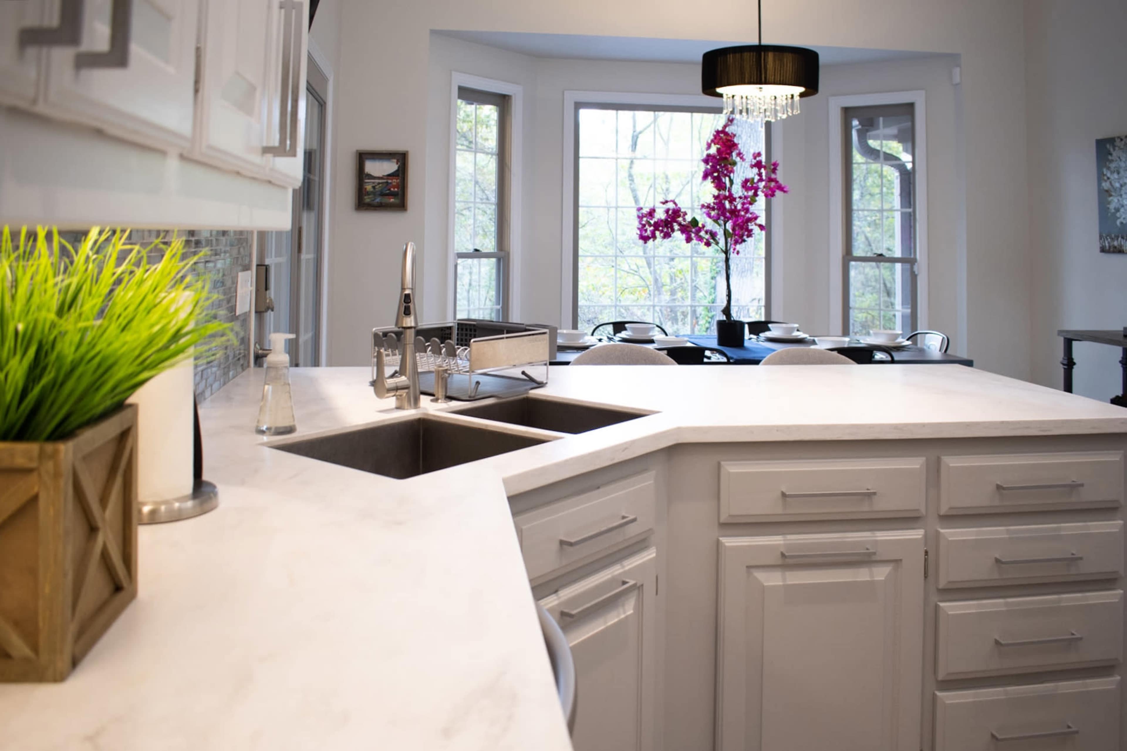 A modern kitchen features white cabinets, a marble countertop with a double sink, and a bay window with a view of a dining area adorned with colorful flowers.