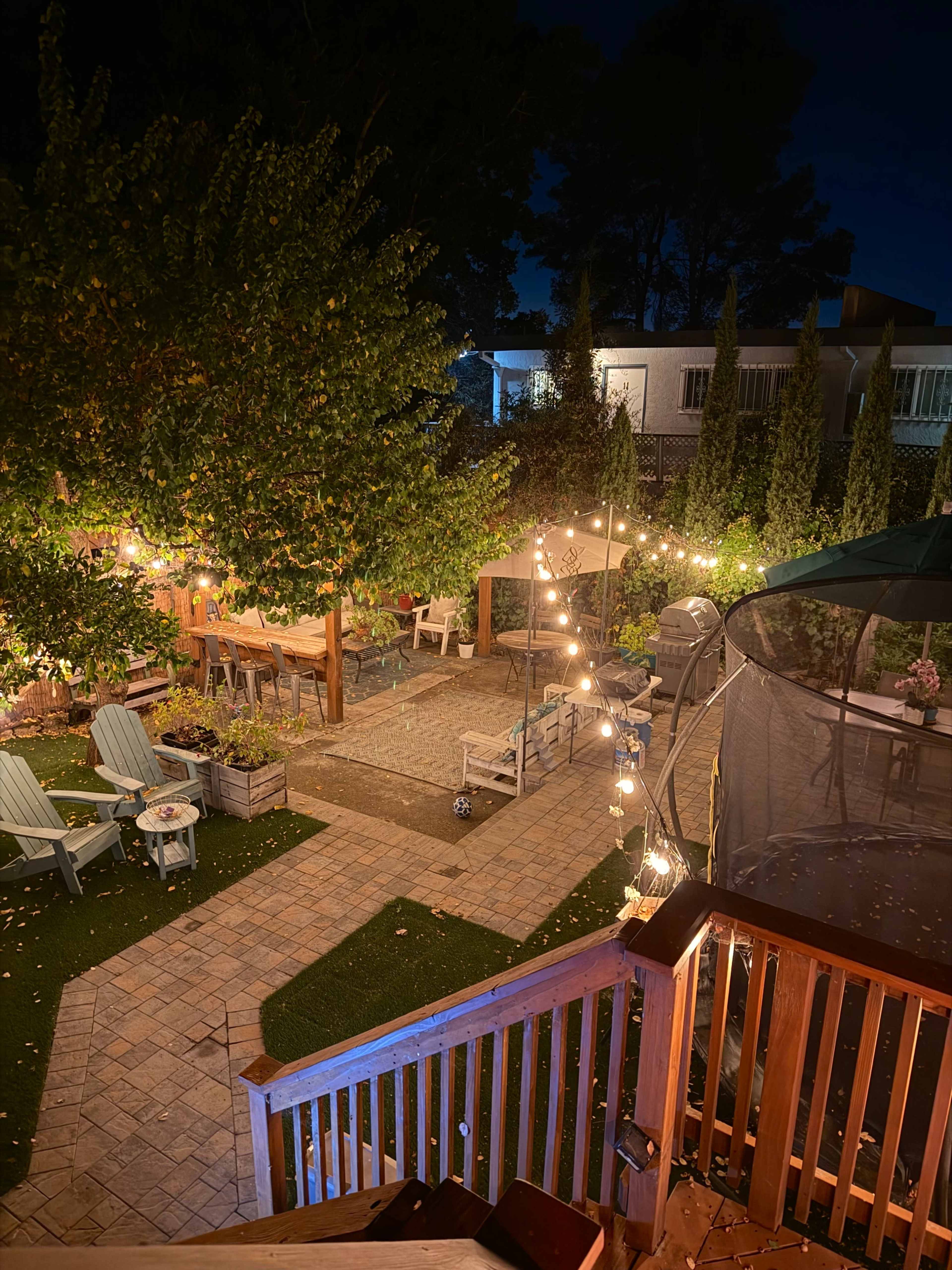 The image shows a well-lit backyard with a patio area containing string lights, seating, and landscaping features, set against a darkening sky.