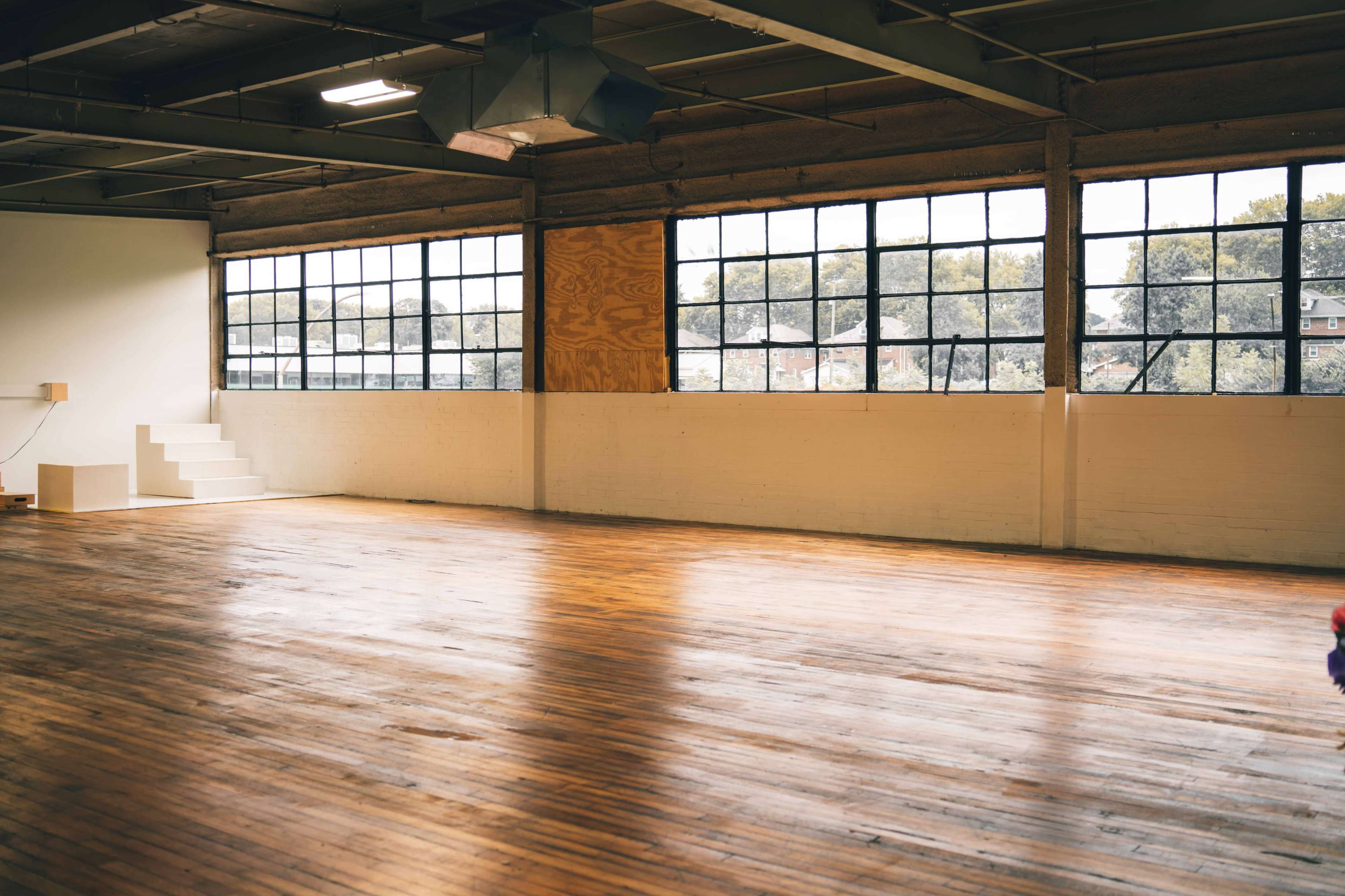 The image shows a spacious, industrial-style room with large windows, wooden floors, and simple white walls, featuring a staircase in the background.