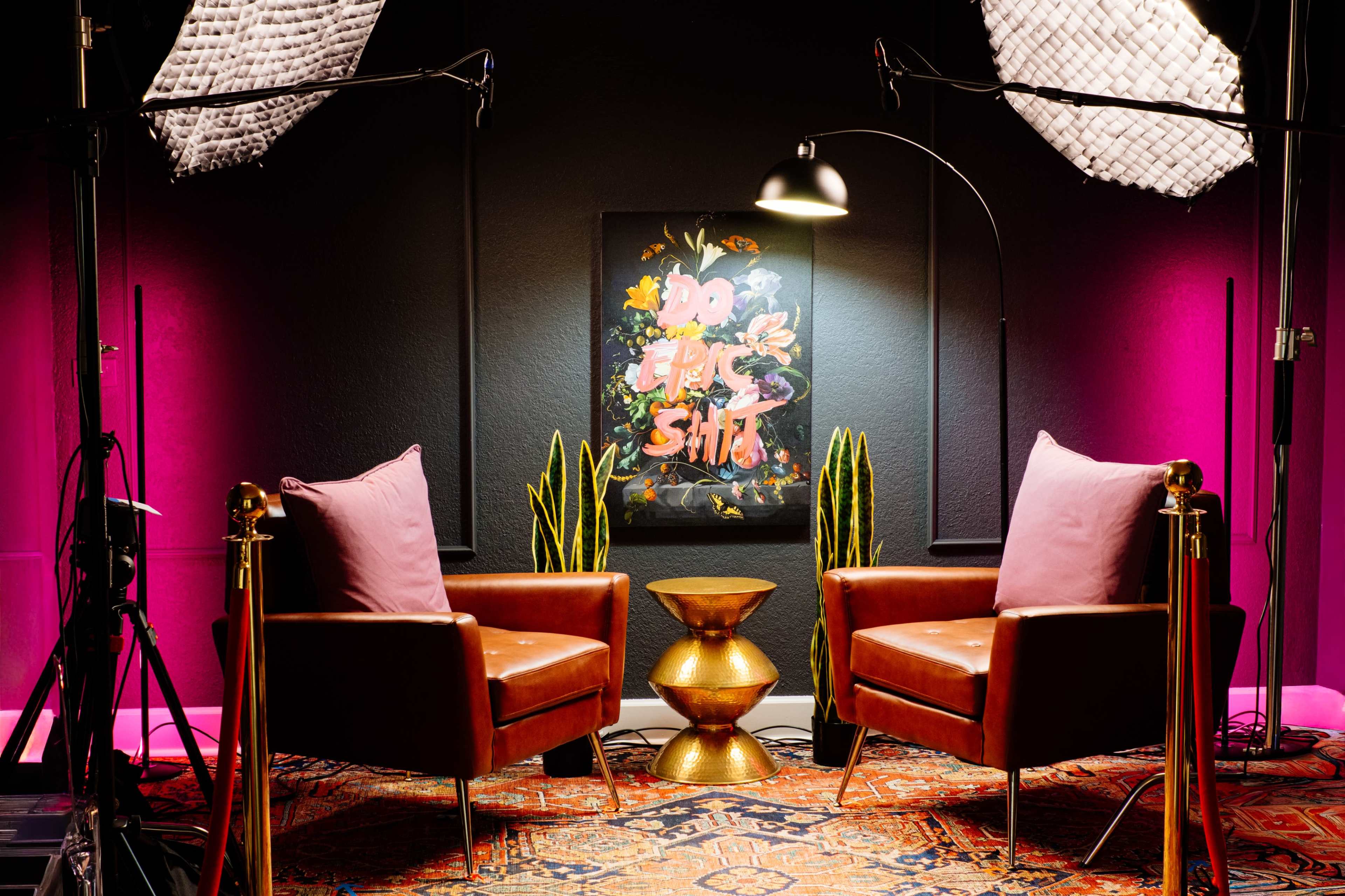 The image features a modern, styled interview setup with two brown leather chairs facing a colorful wall art piece, illuminated by soft lighting and surrounded by potted plants.