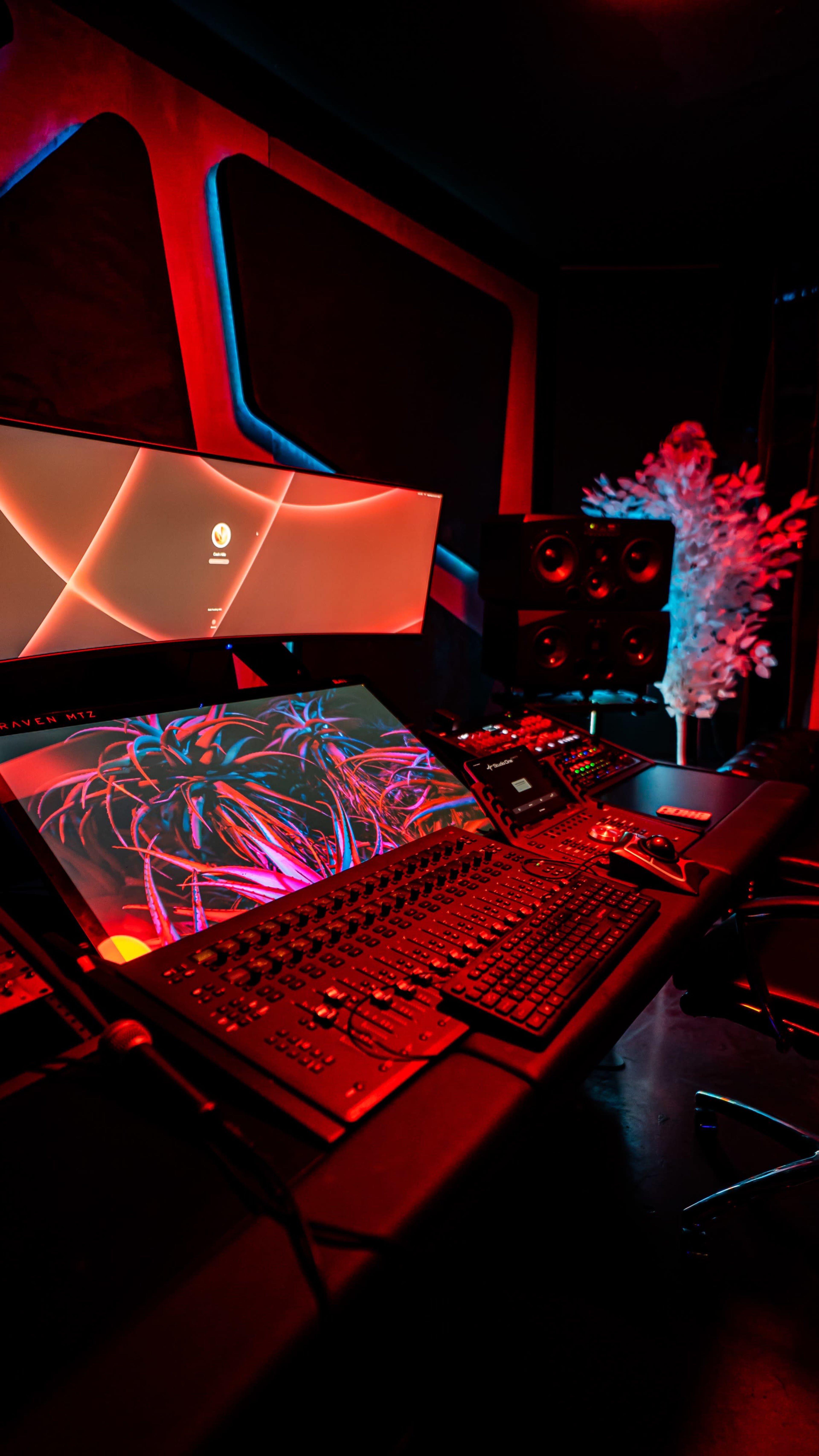 A modern music studio features a large digital mixing console, dual computer monitors, and colorful ambient lighting.