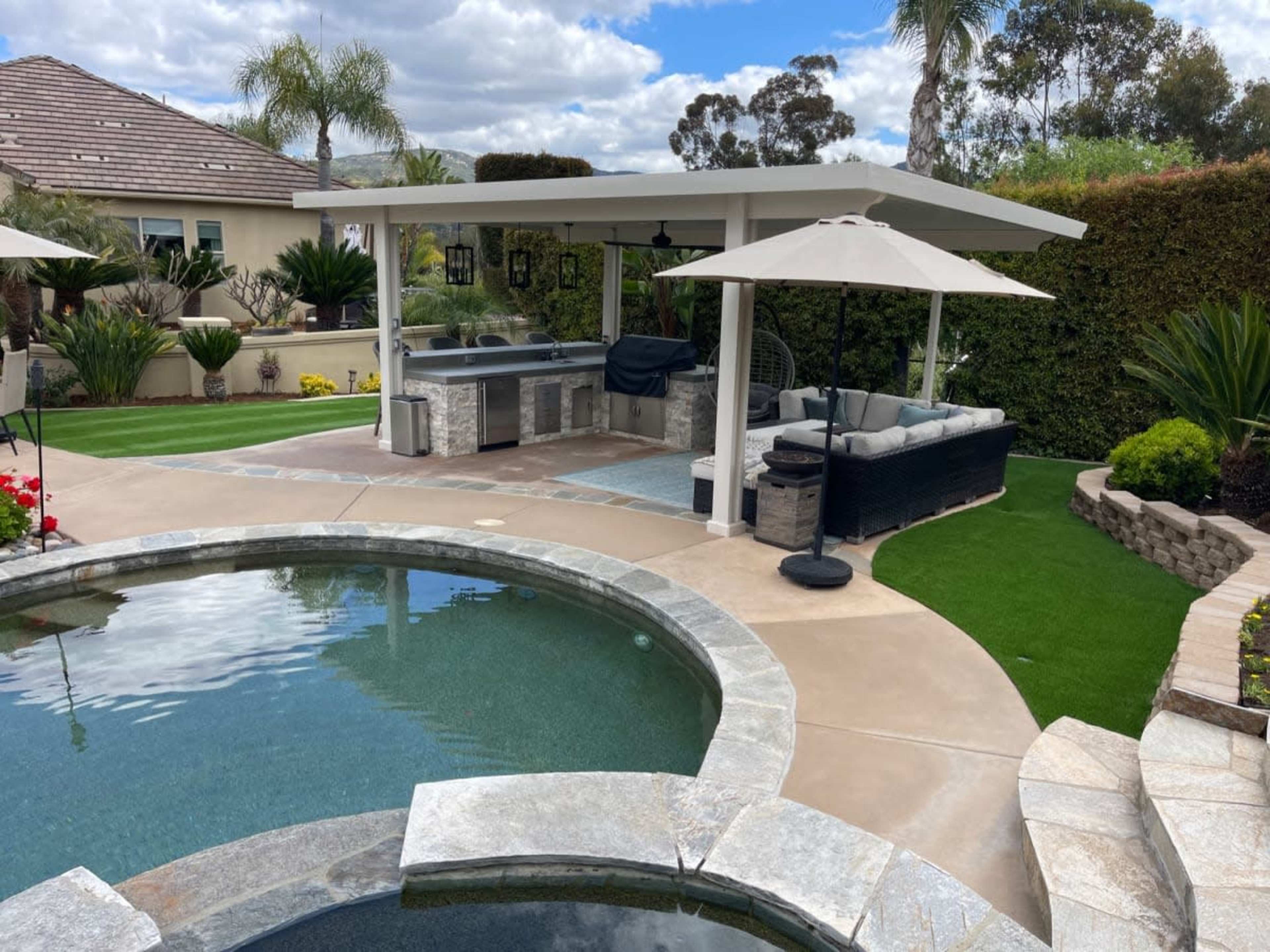 The image shows a backyard with a pool, a spa area, and a covered seating space featuring lounge furniture and an umbrella.