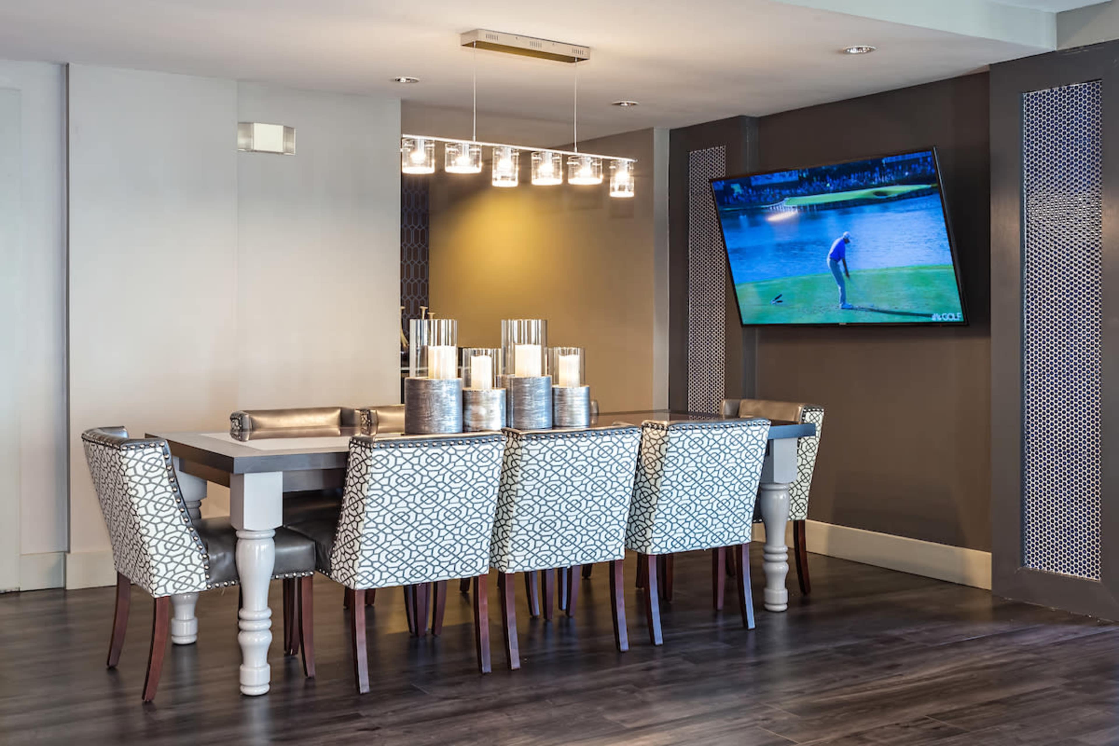 A modern dining room features a long table surrounded by patterned chairs and a large TV mounted on the wall.