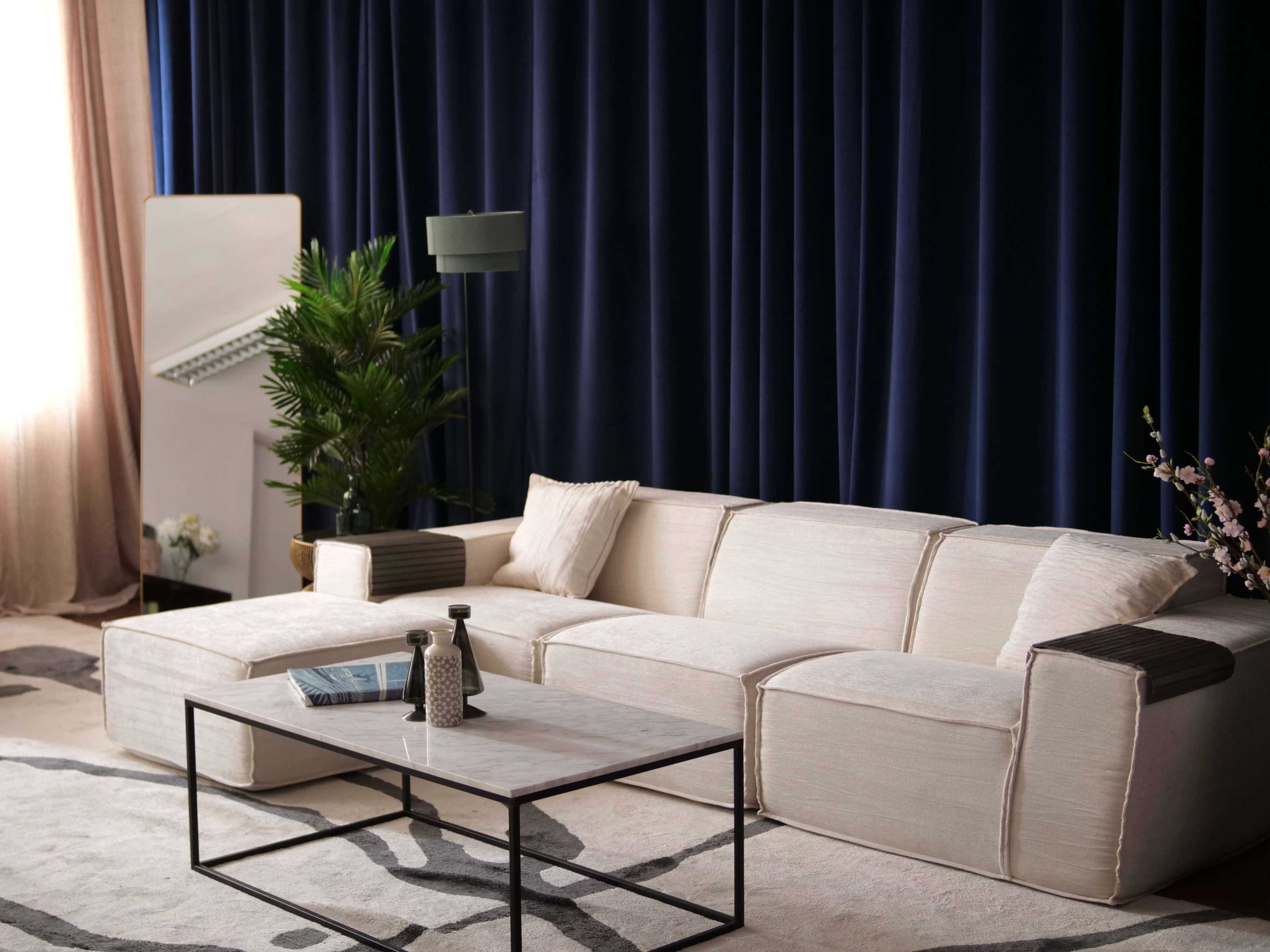 A modern living room features a light-colored sectional sofa, a coffee table, and a tall plant against dark blue curtains.