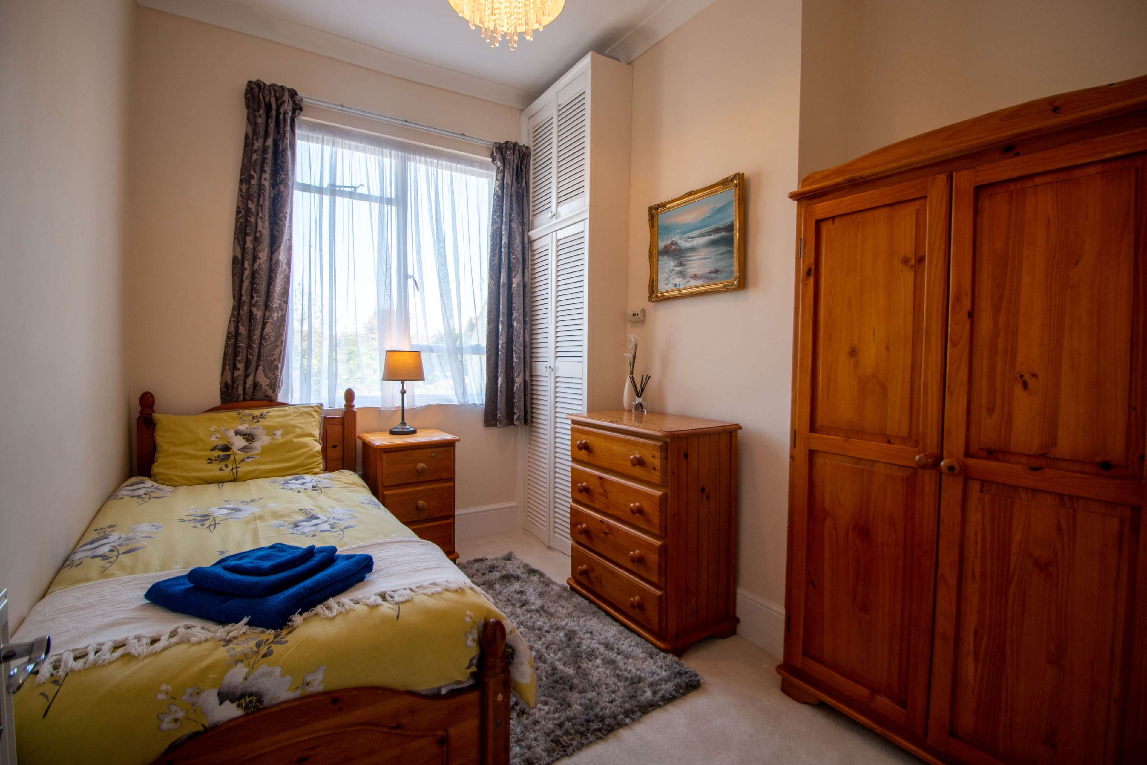 The image shows a small bedroom featuring a single bed with floral bedding, a bedside lamp, a chest of drawers, and a wooden wardrobe.