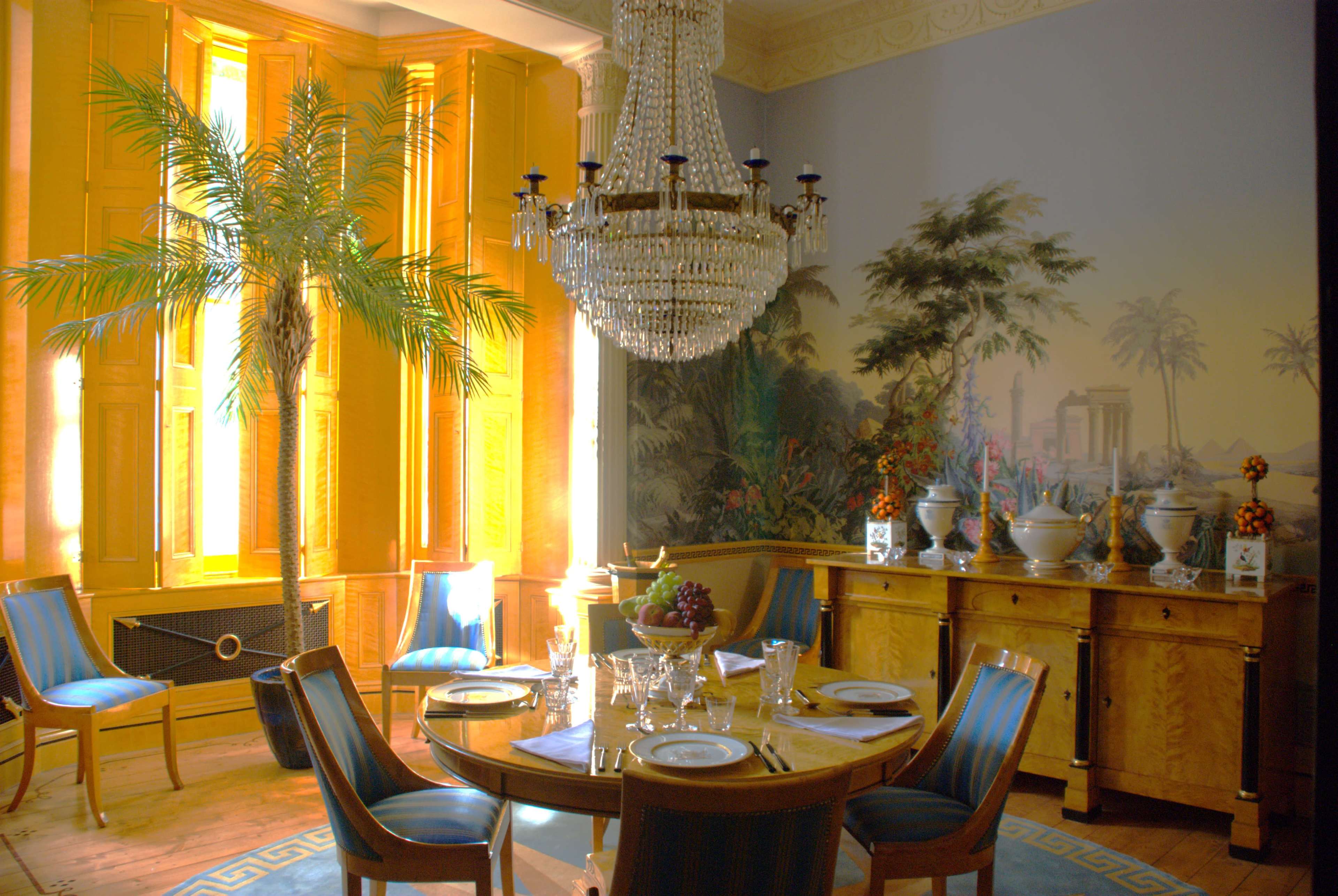 The image features a dining room with a round table set with plates and glassware, surrounded by chairs, a large chandelier overhead, and a palm tree in the corner.
