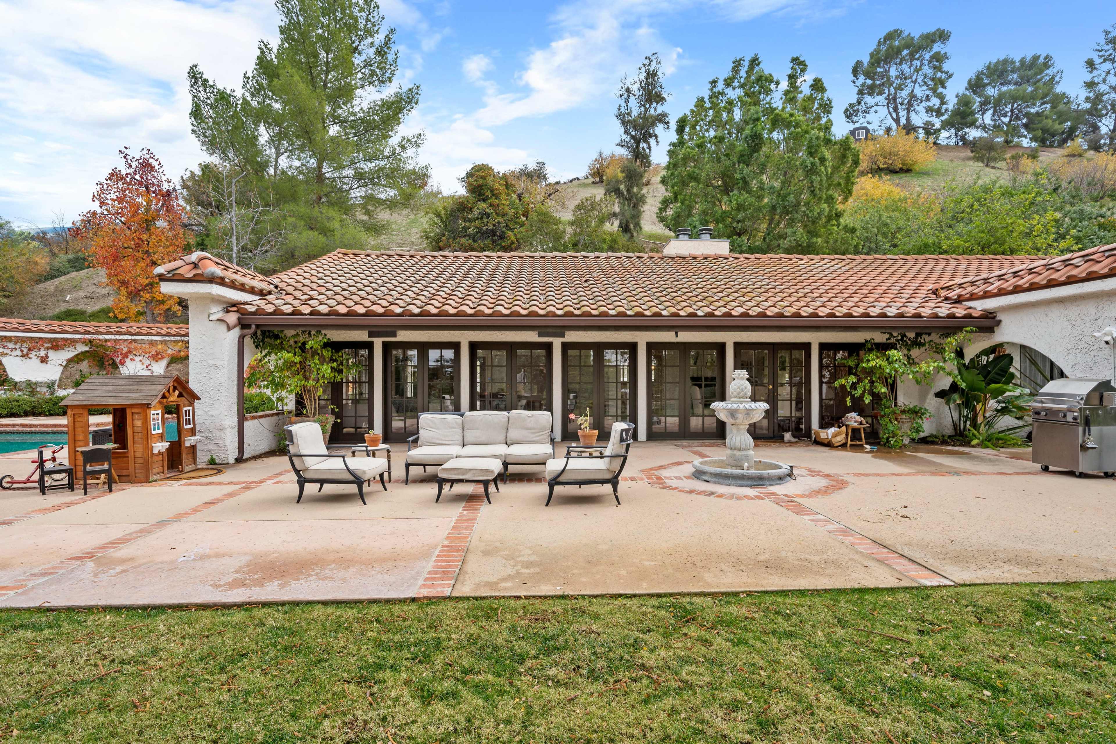 A patio area features outdoor seating and a fountain, surrounded by lush landscaping and a view of a hillside.