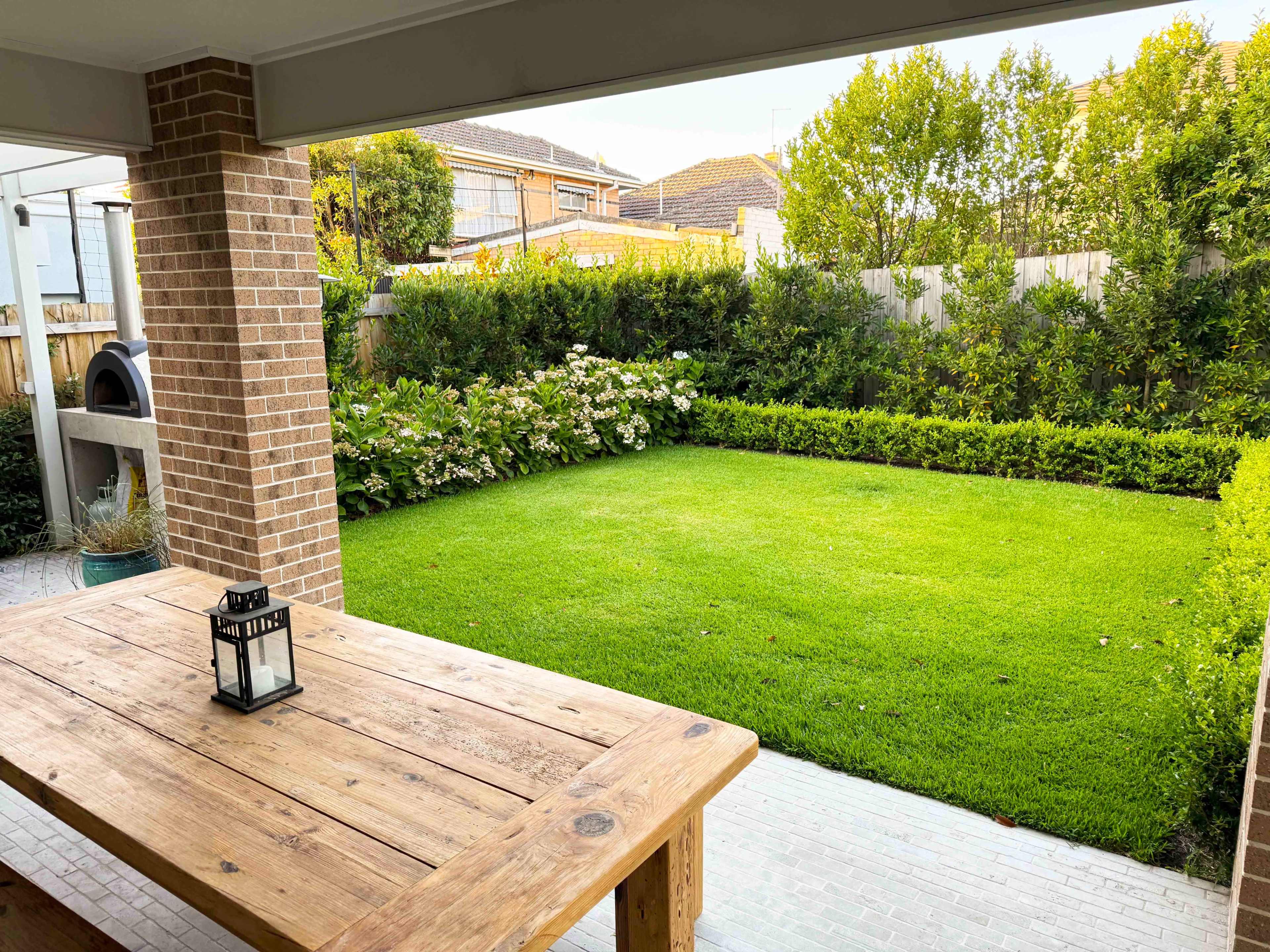 The image shows a well-maintained backyard with a neatly trimmed lawn, surrounded by lush greenery and a patio area featuring a wooden table and lantern.