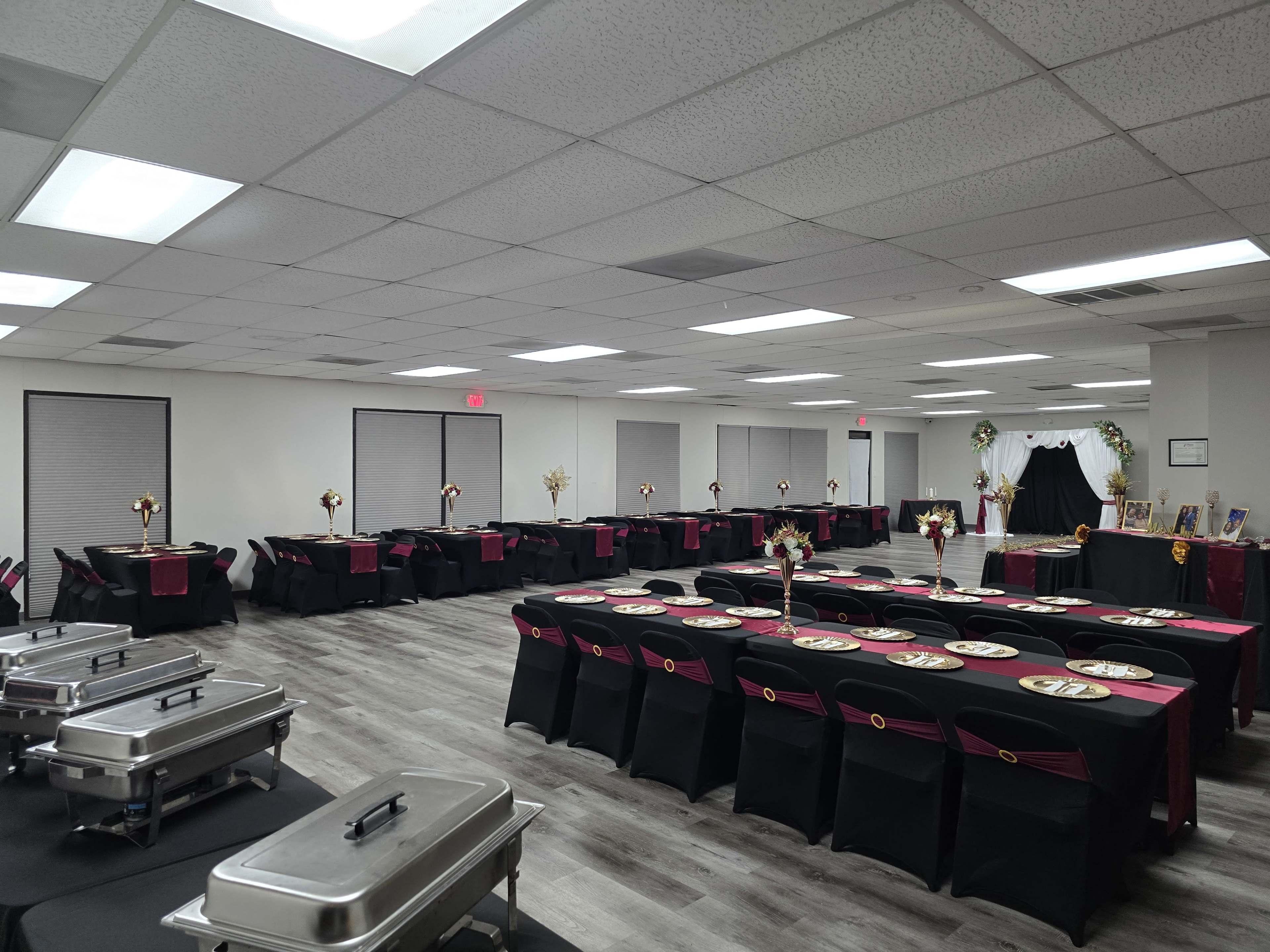 The image shows a banquet hall arranged with several long tables covered in black tablecloths and accented with burgundy, set up for an event, with food warming trays placed on one side of the room.