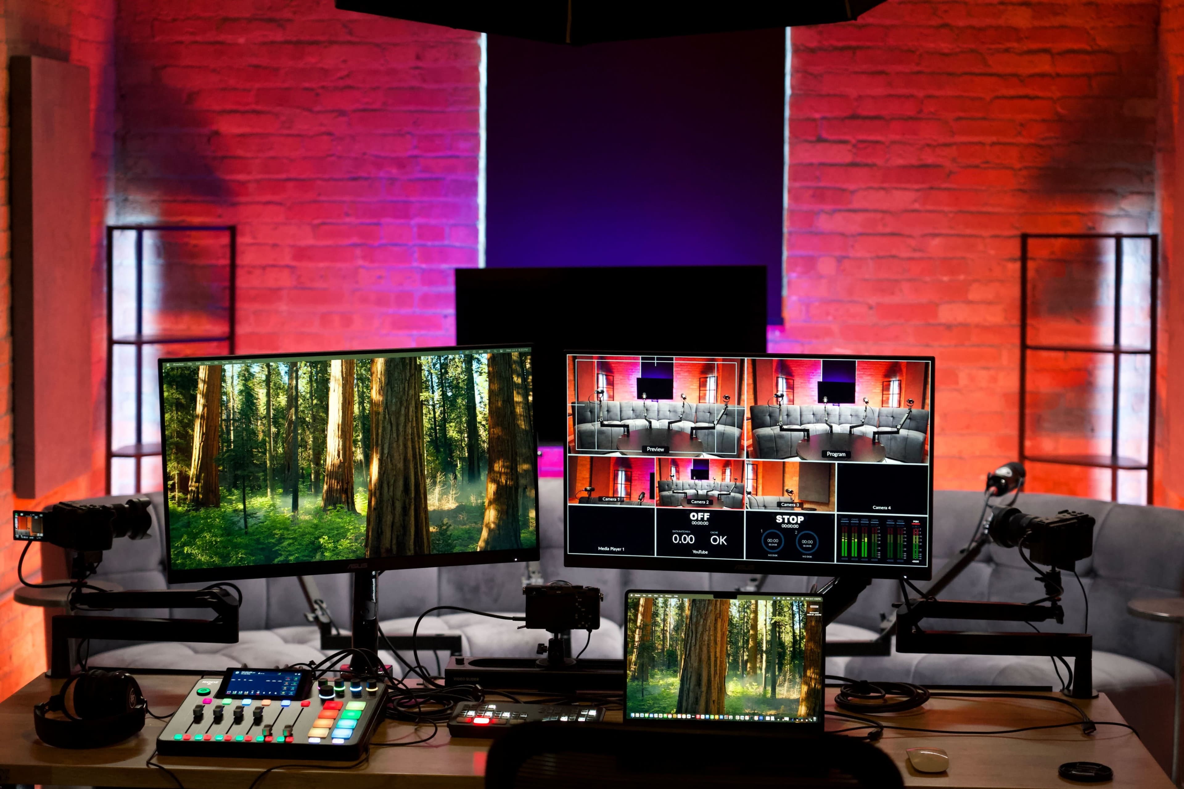 A workspace featuring multiple monitors and a control surface, illuminated by colorful lighting against a brick wall backdrop.