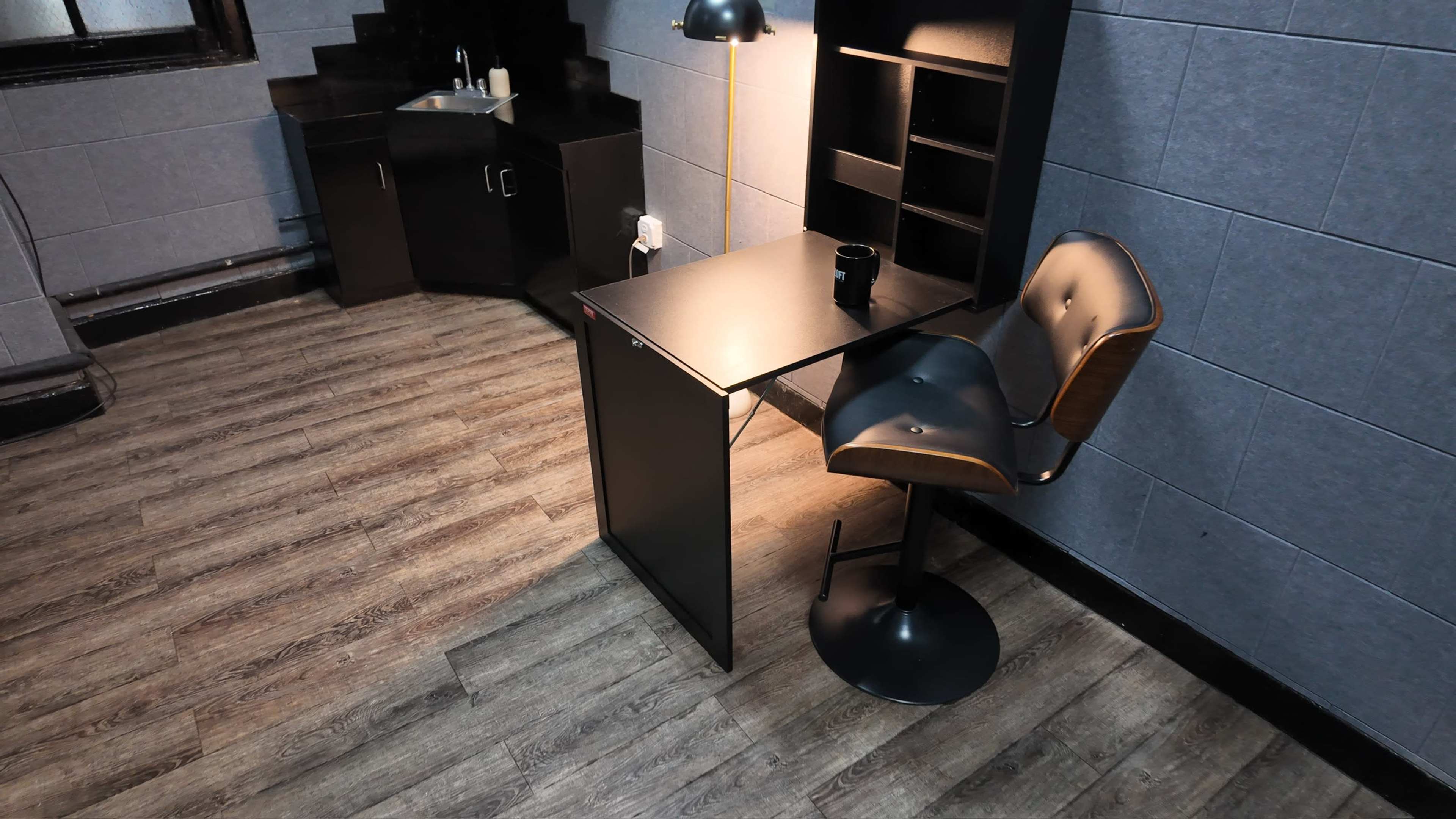 The image shows a small, minimalistic workspace featuring a desk and a high-backed stool, with a dark cabinet and a light illuminating the area.