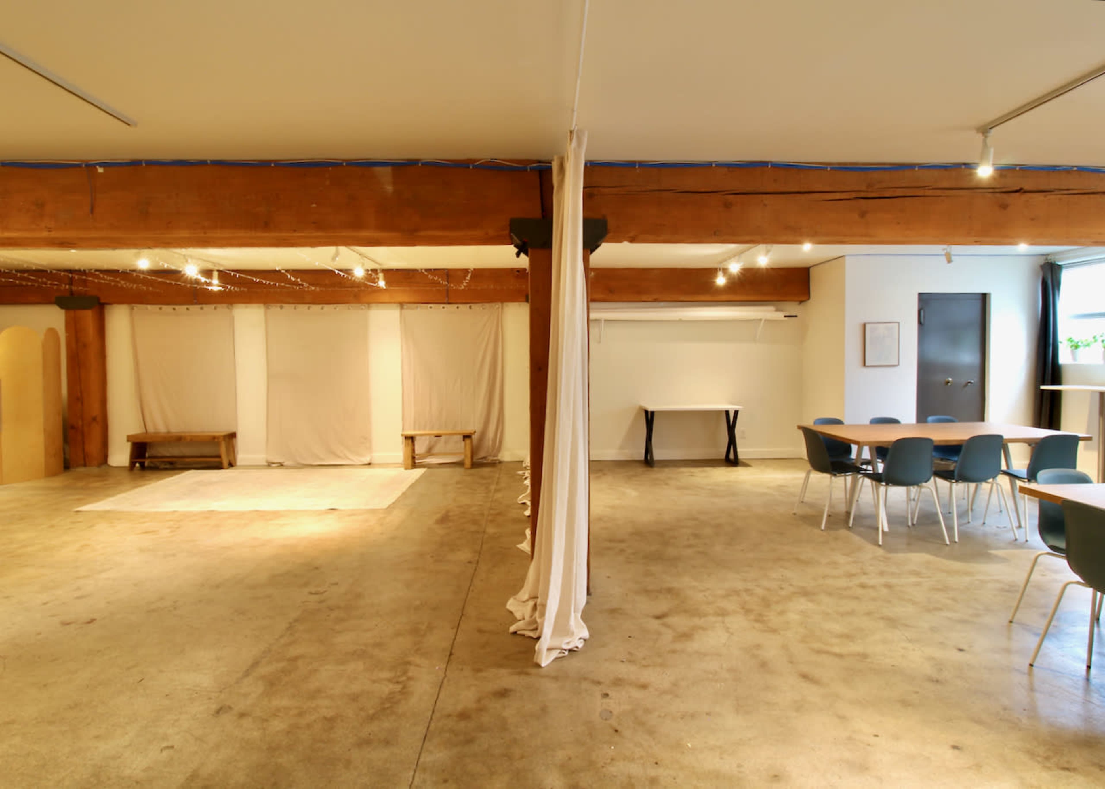 The image shows a spacious, well-lit interior with exposed wooden beams, a concrete floor, and a few tables arranged for seating.