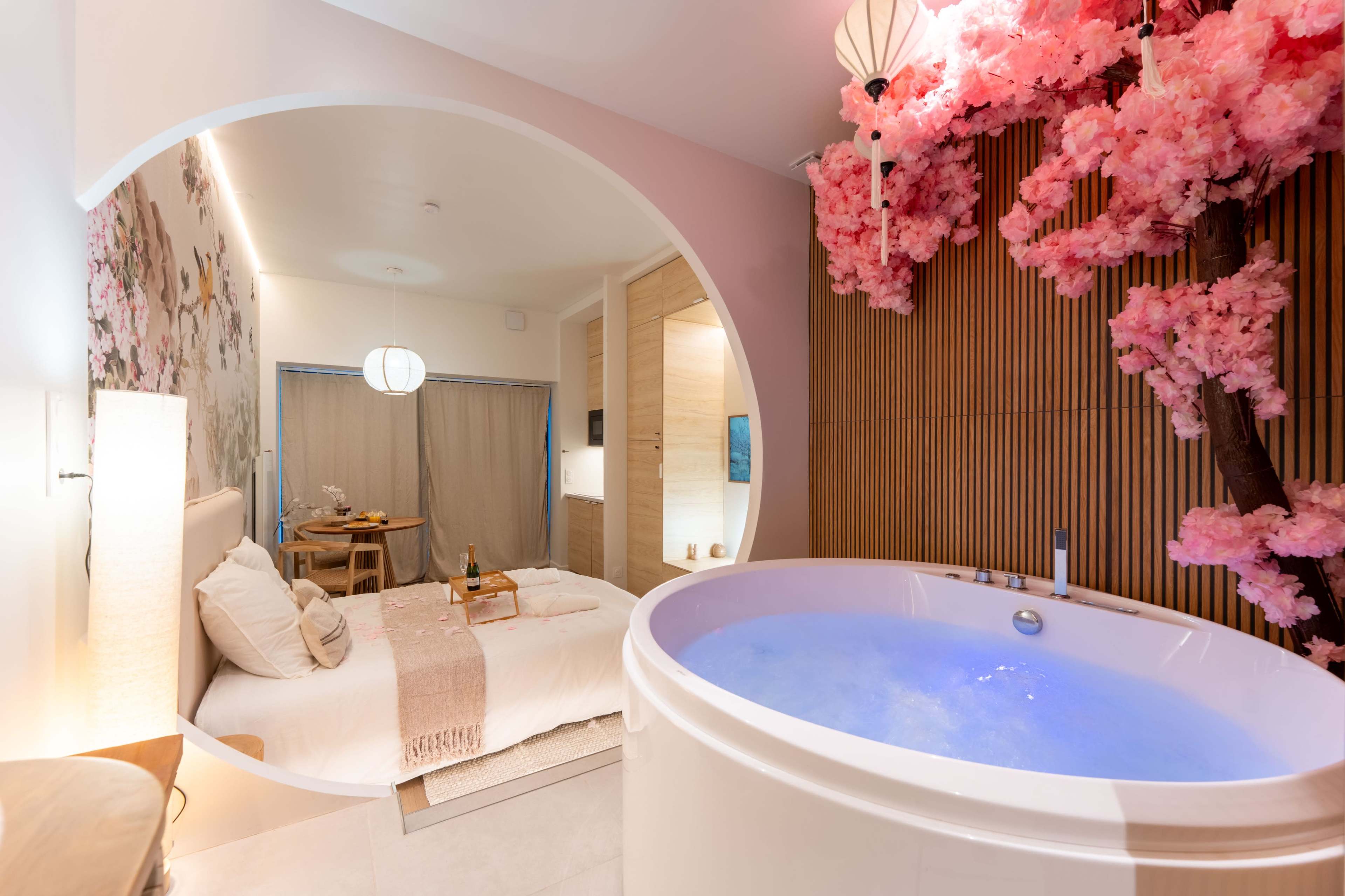 The image shows a modern hotel room featuring a round view into a bathroom area with a circular bathtub illuminated in blue, surrounded by pink cherry blossom decorations.