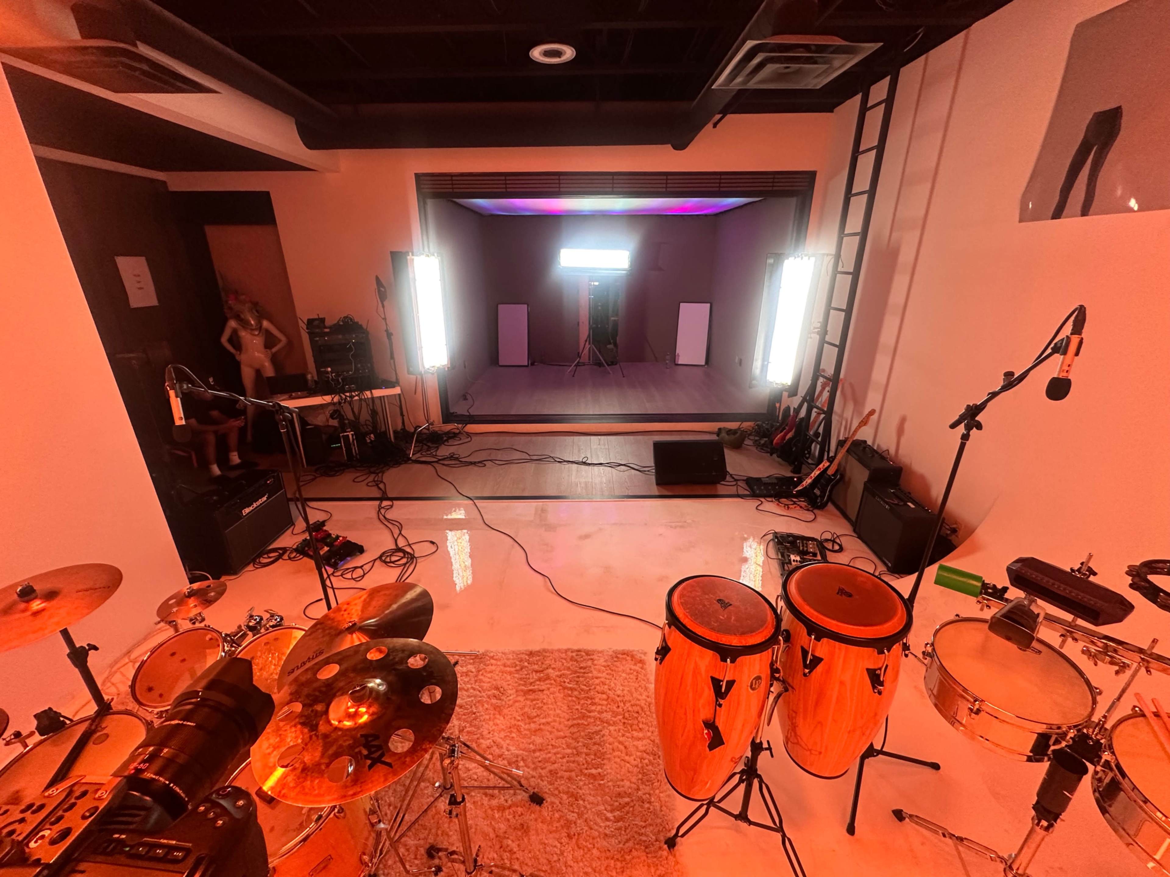 The image shows a music studio setup with various instruments, including drums and congas, along with multiple lighting setups and microphones arranged on the floor.