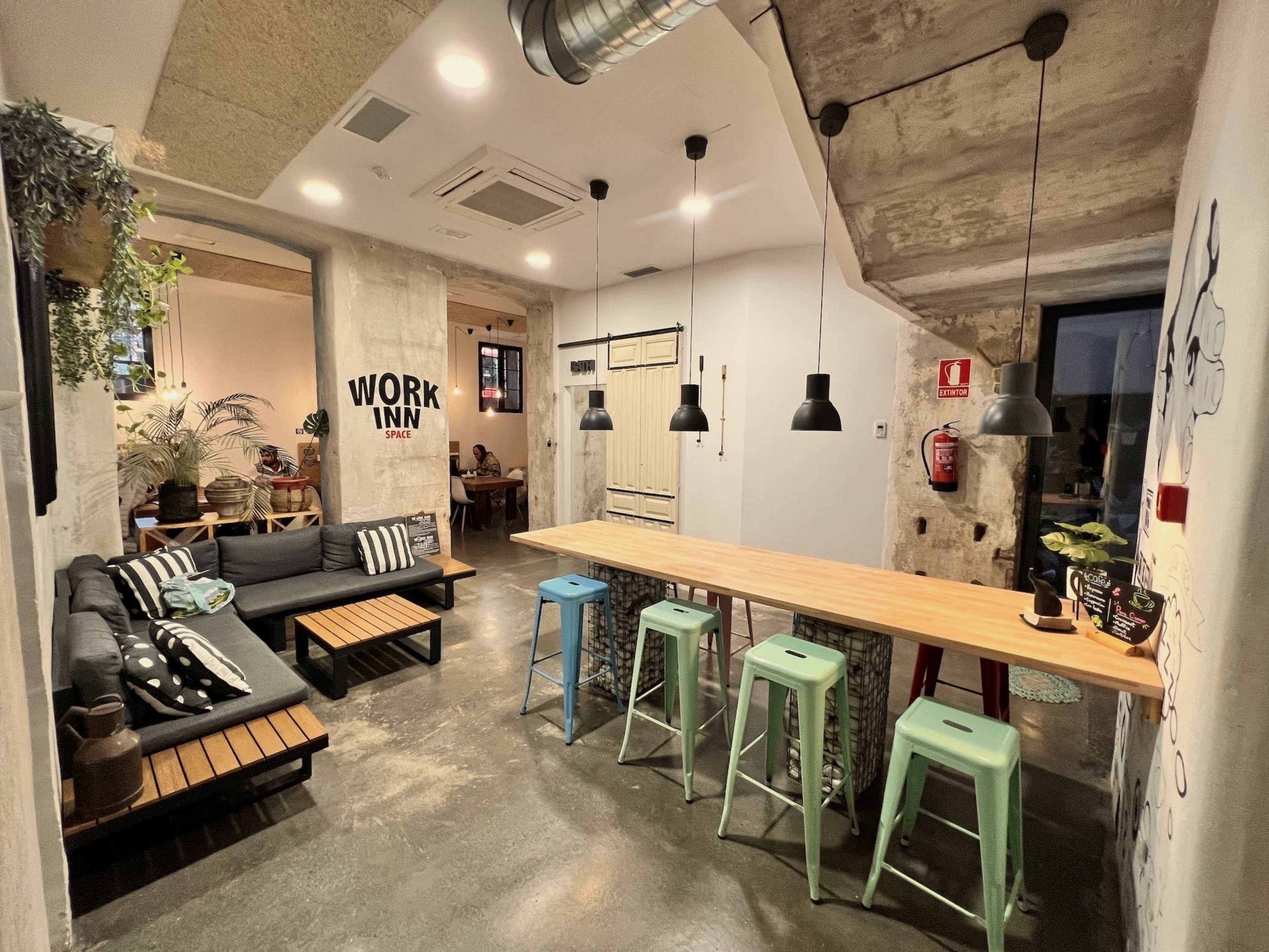 The image shows a modern co-working space with a seating area featuring a couch, a wooden table with colorful stools, and a wall displaying the text "WORK INN."