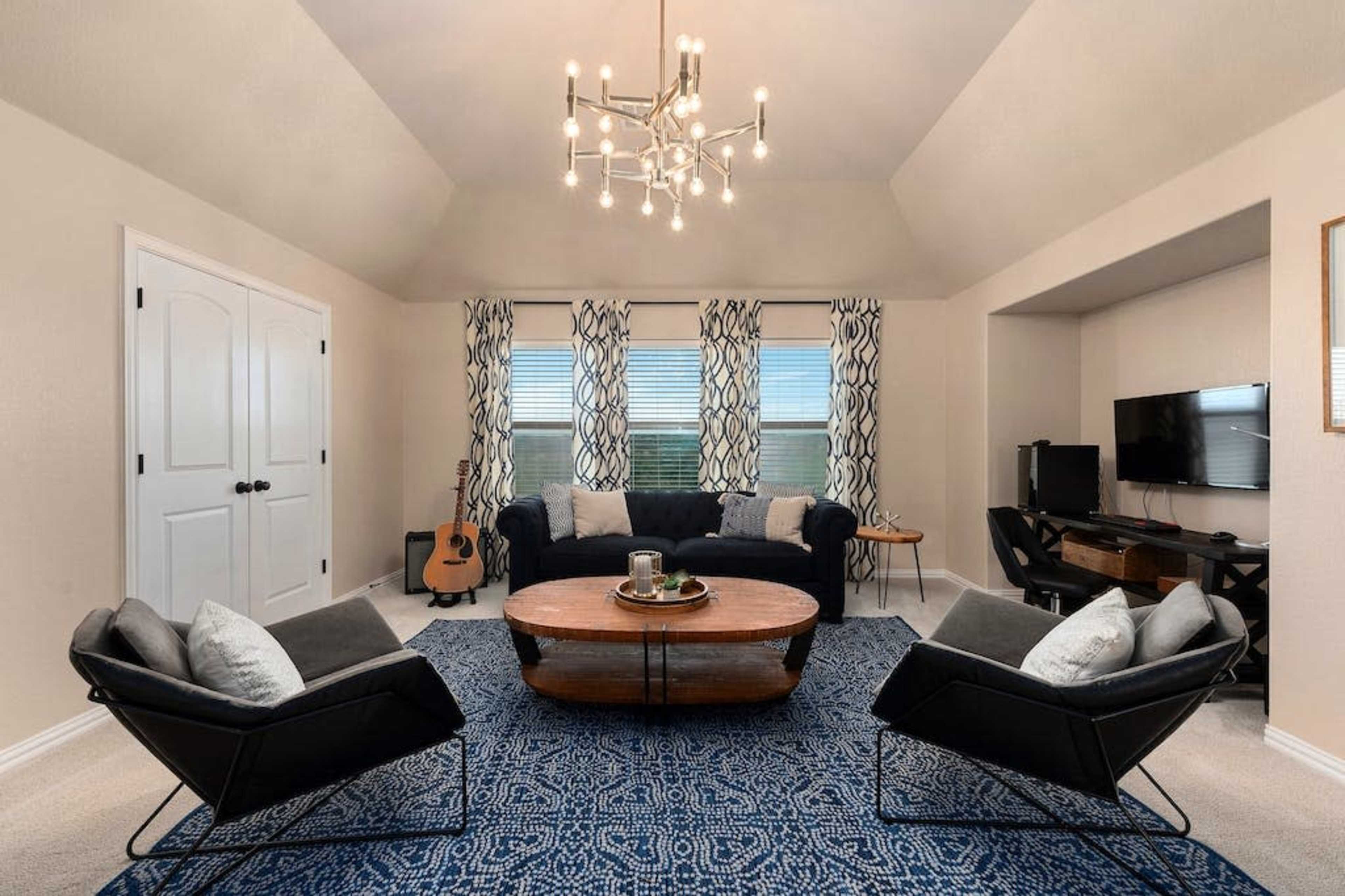A cozy living room with a blue patterned rug, a large circular coffee table, two armchairs, and a couch, set against windows with patterned curtains and a TV console.