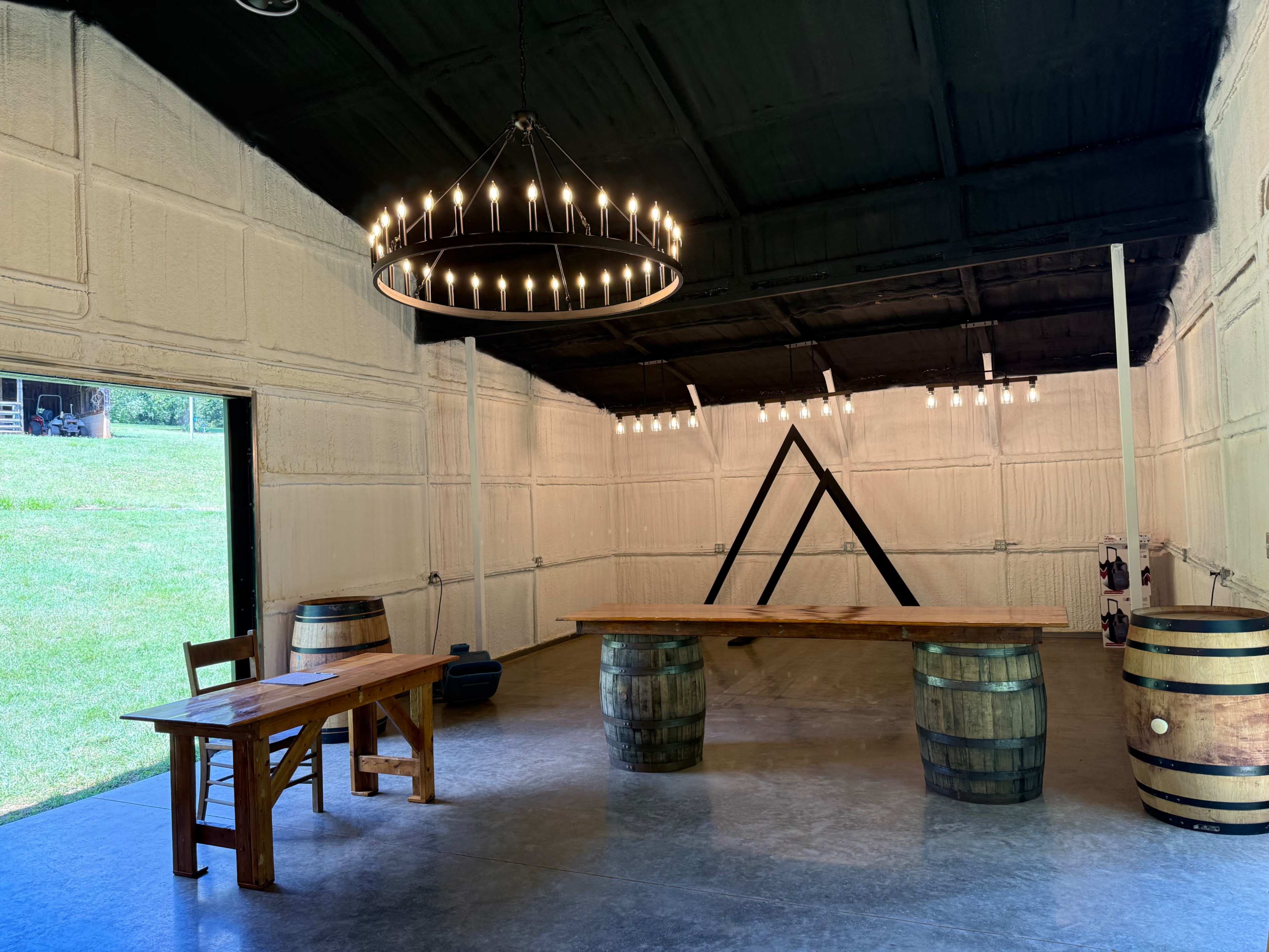 The image shows a spacious room with a wooden table and chairs, two large barrels, and a chandelier hanging from the ceiling, all situated against a backdrop of white walls and a view of grassy outdoors.