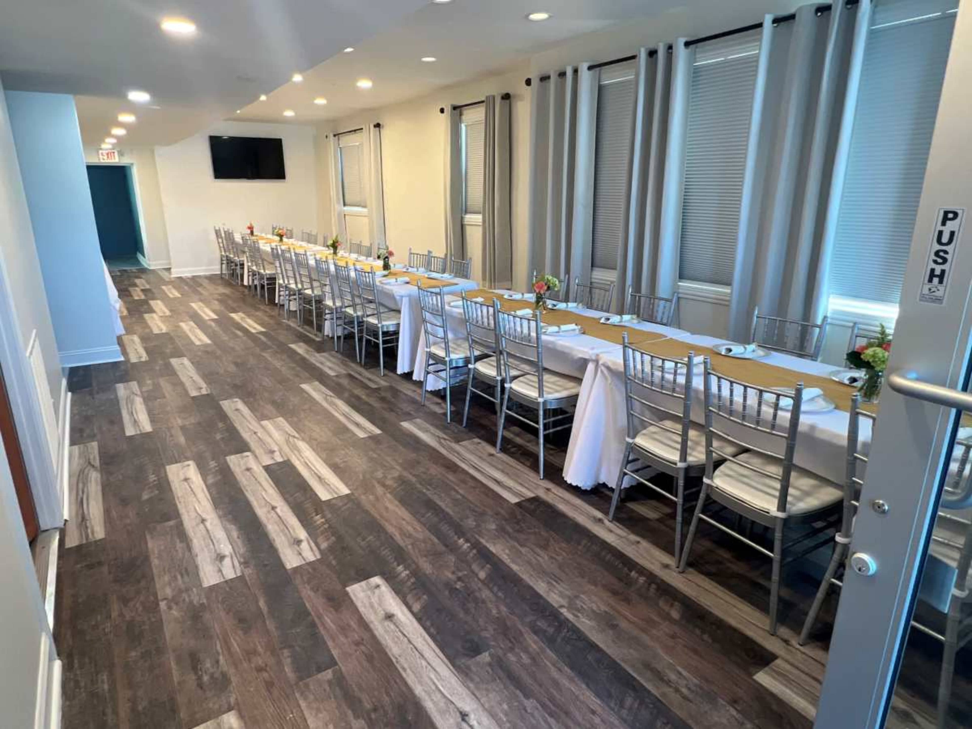 The image shows a long, rectangular dining setup with a table draped in a white tablecloth and adorned with small centerpieces, surrounded by silver chiavari chairs in a well-lit interior.
