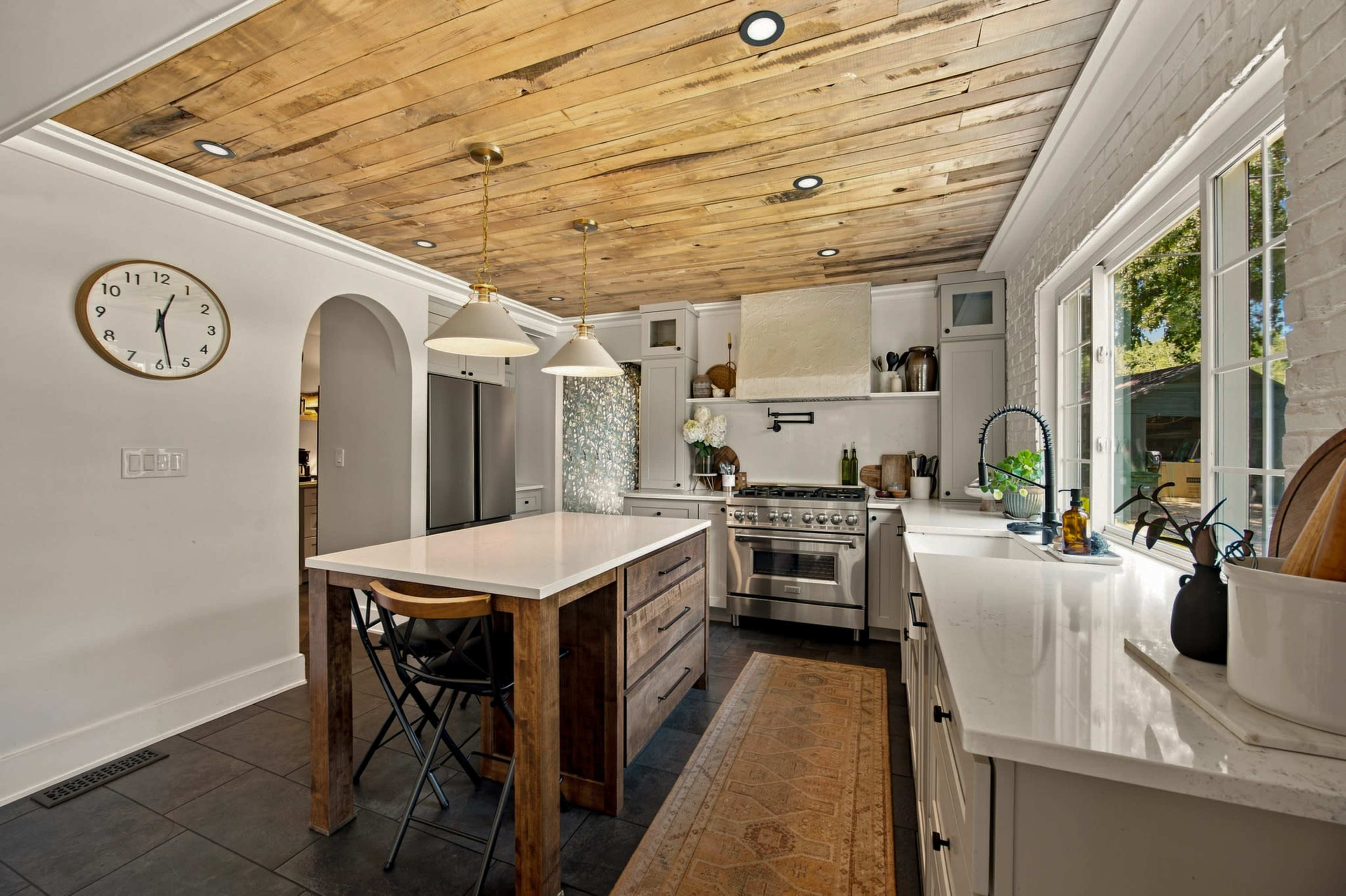 The kitchen features a central island with wooden base, white cabinets, a stainless steel stove, and large windows that provide natural light.