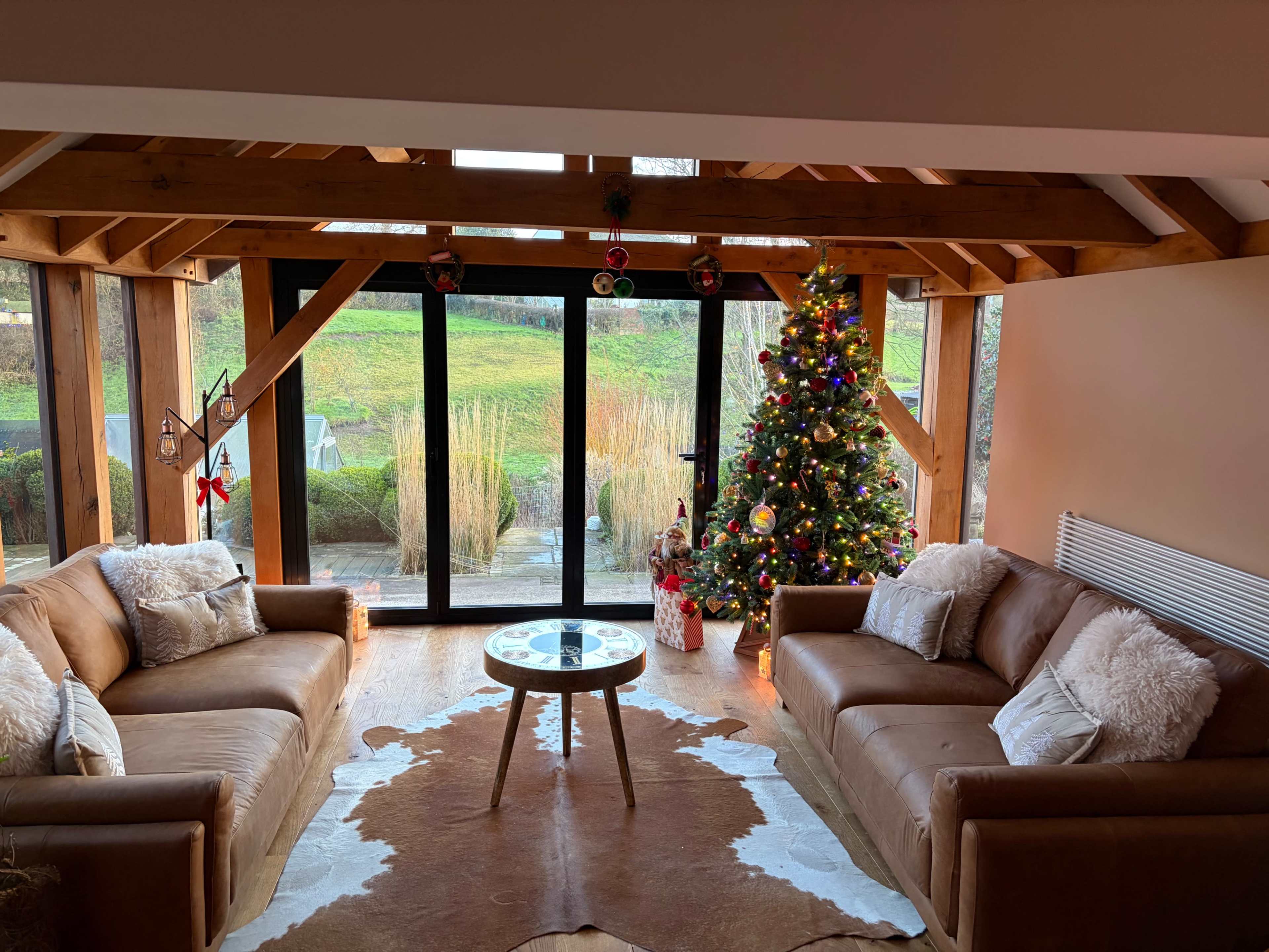 The image features a cozy living room with wooden beams, two brown sofas, a round glass coffee table, and a decorated Christmas tree next to large sliding glass doors that open to a view of greenery outside.