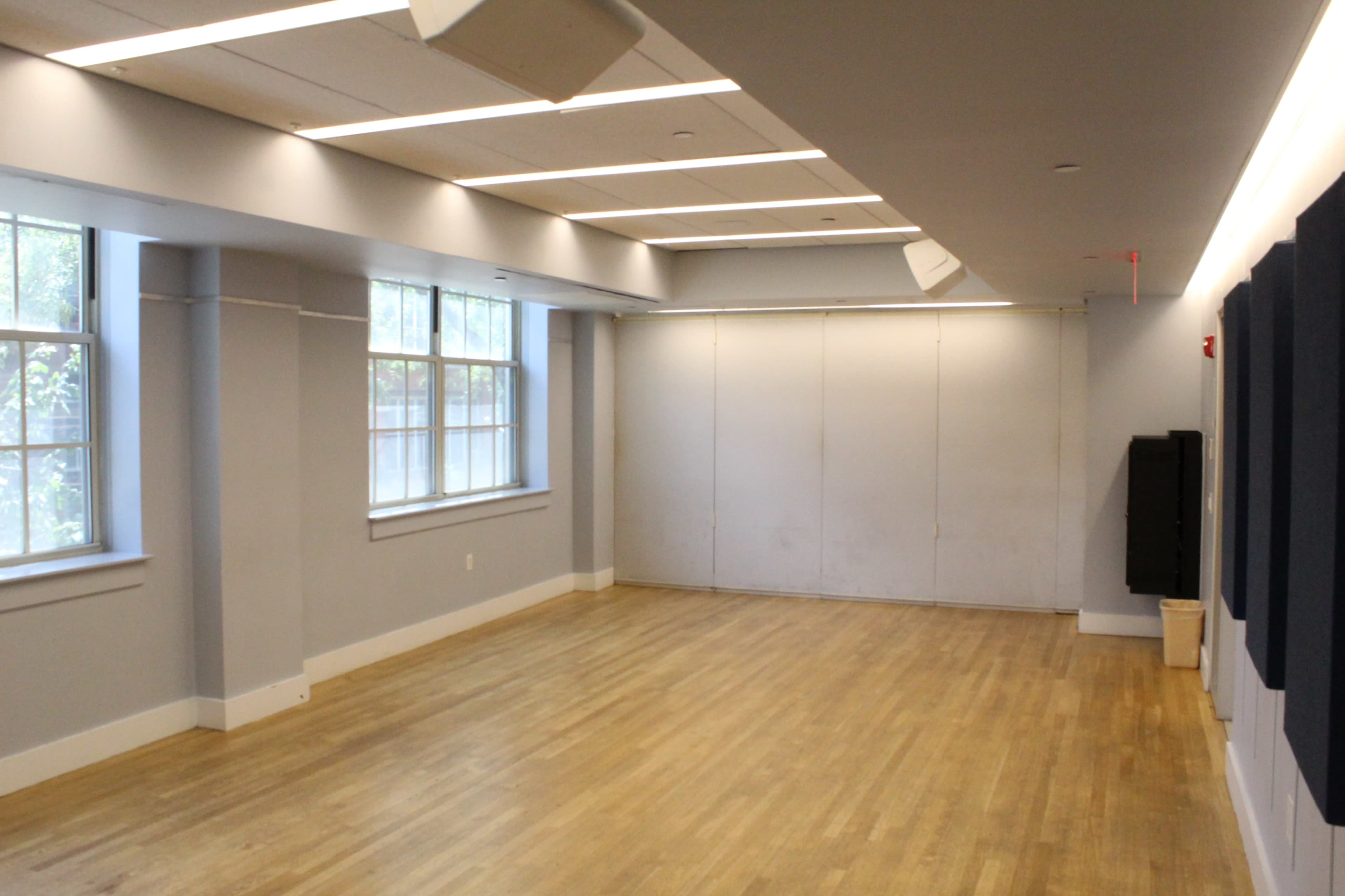 The image shows a spacious, brightly lit room with large windows and wooden flooring, featuring a blank wall and some soundproofing panels.