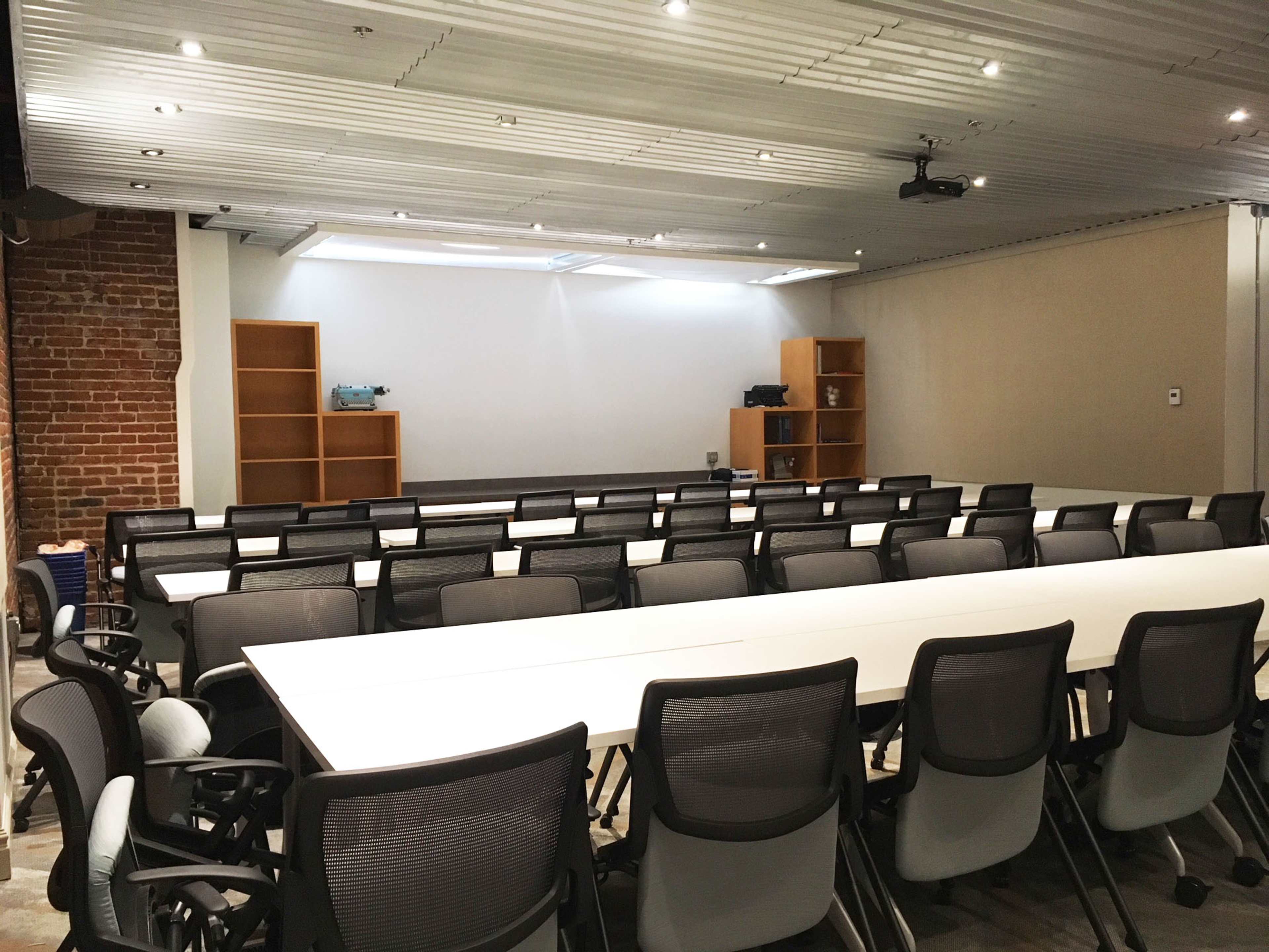 A meeting room features rows of chairs and long tables arranged for presentations, with shelves along one wall and a projector mounted on the ceiling.