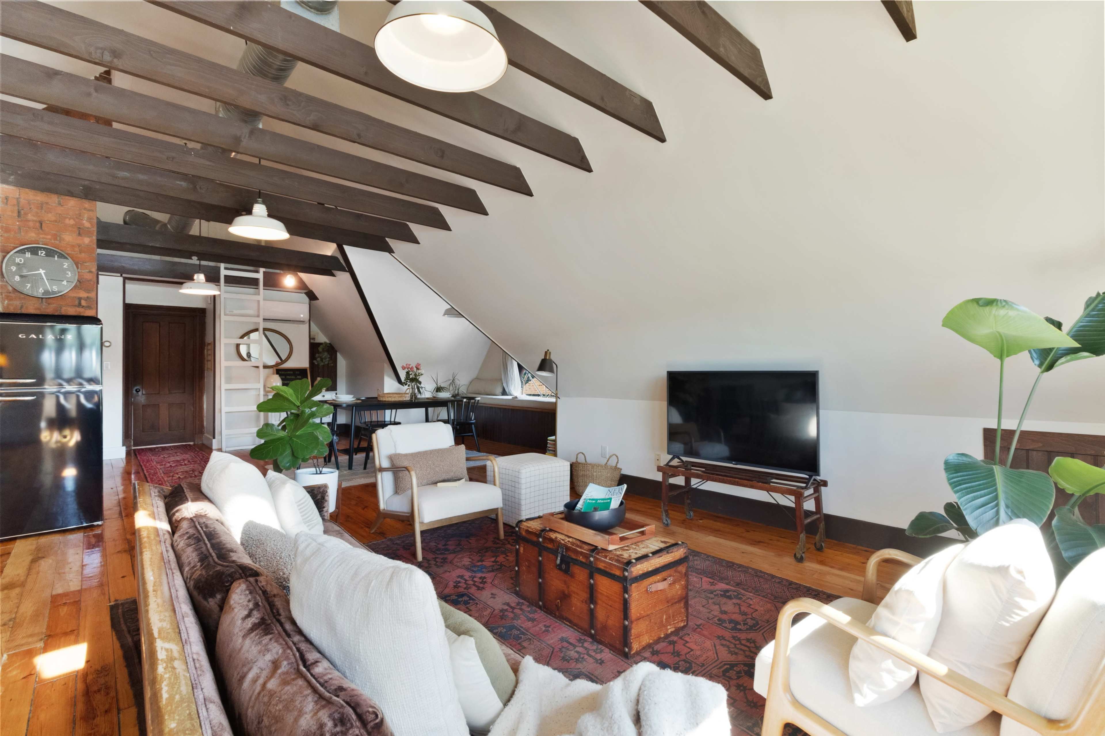 A cozy attic space features wooden beams, a seating area with a sofa and chairs, a television, a vintage trunk, and a small kitchenette area.
