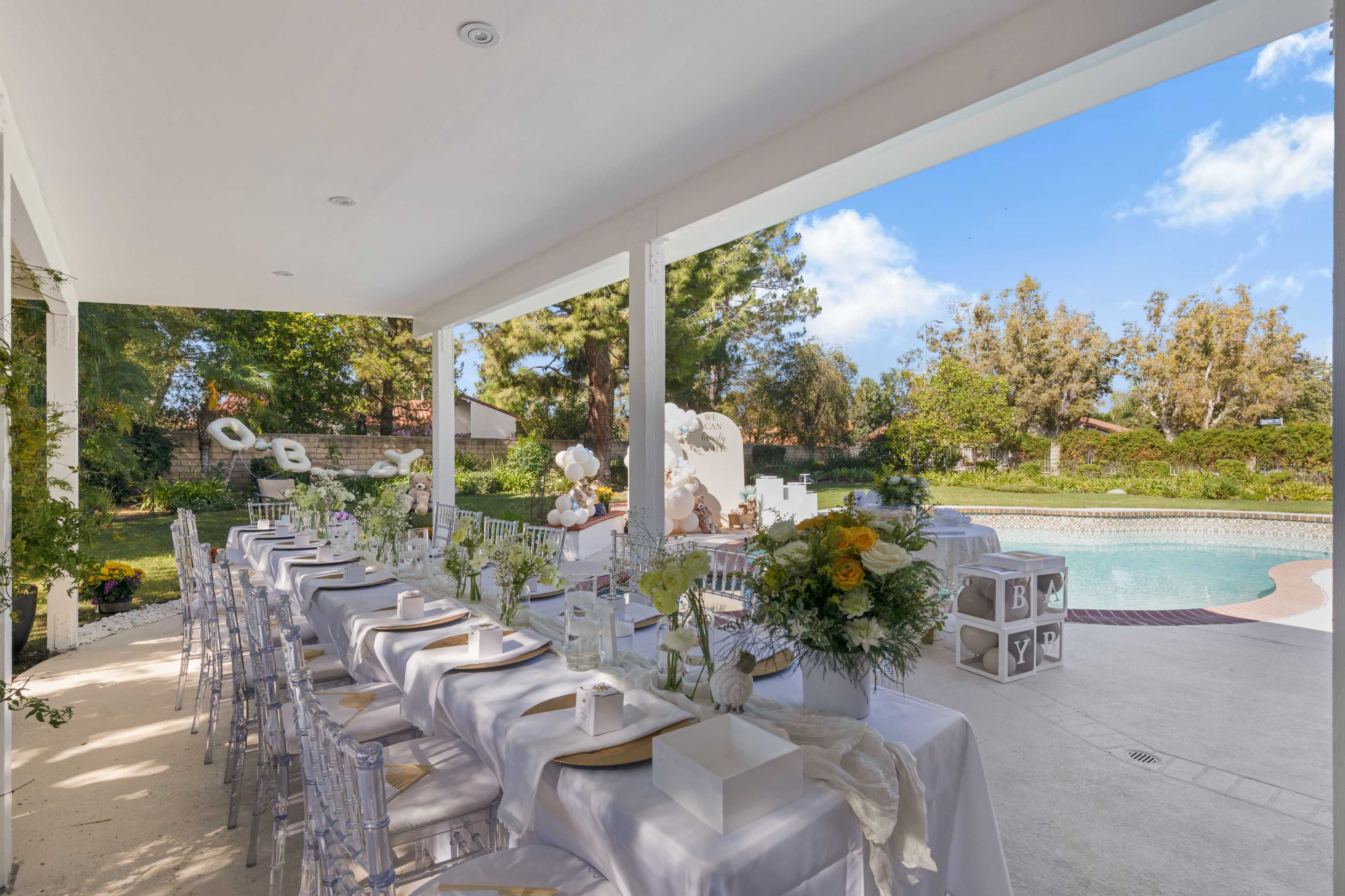 A dining area with elegantly set tables lined under a covered patio overlooks a swimming pool and landscaped garden.