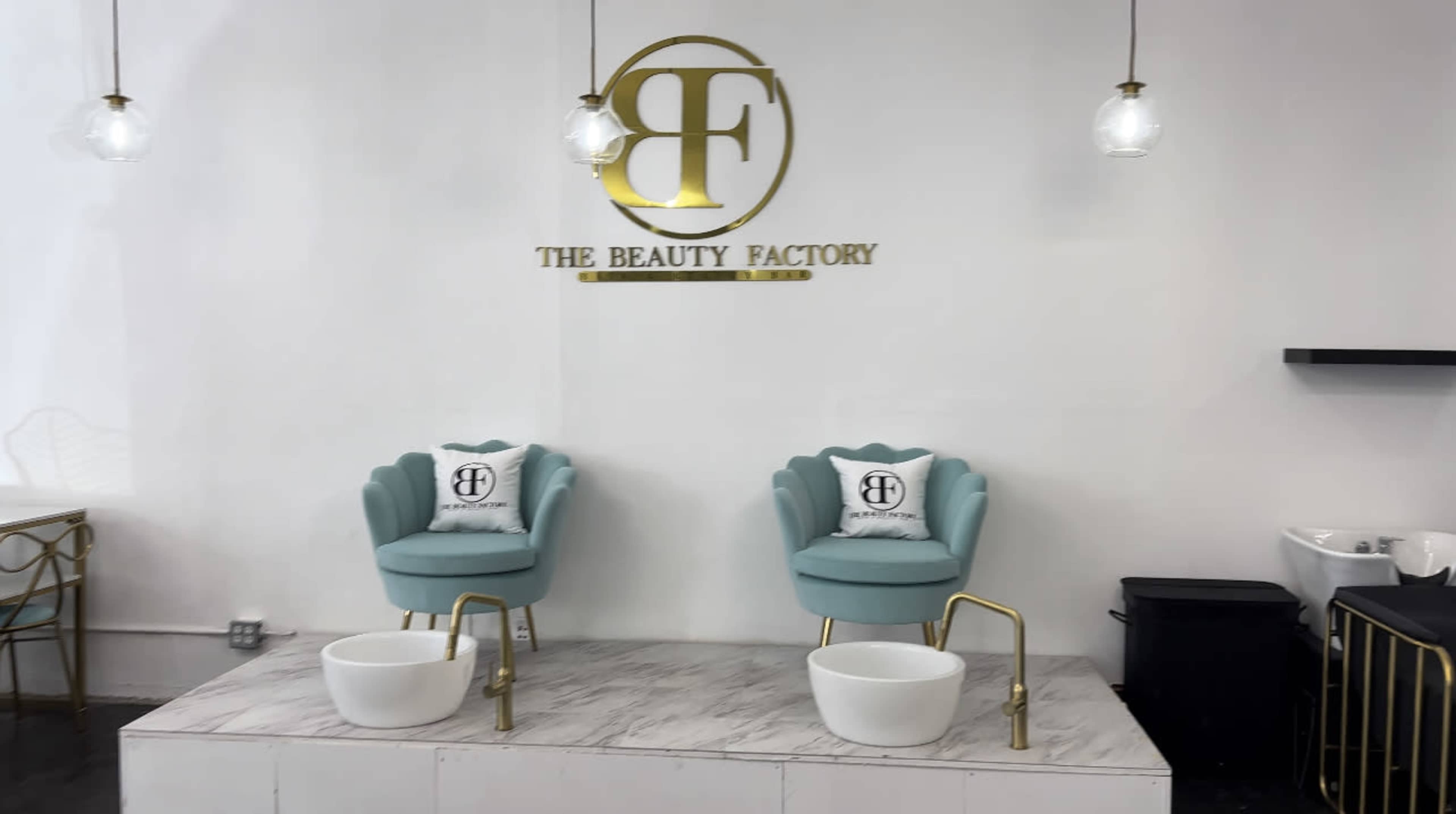 A minimalist beauty salon with two mint green chairs and white foot baths, set against a white wall featuring a gold logo for "The Beauty Factory."