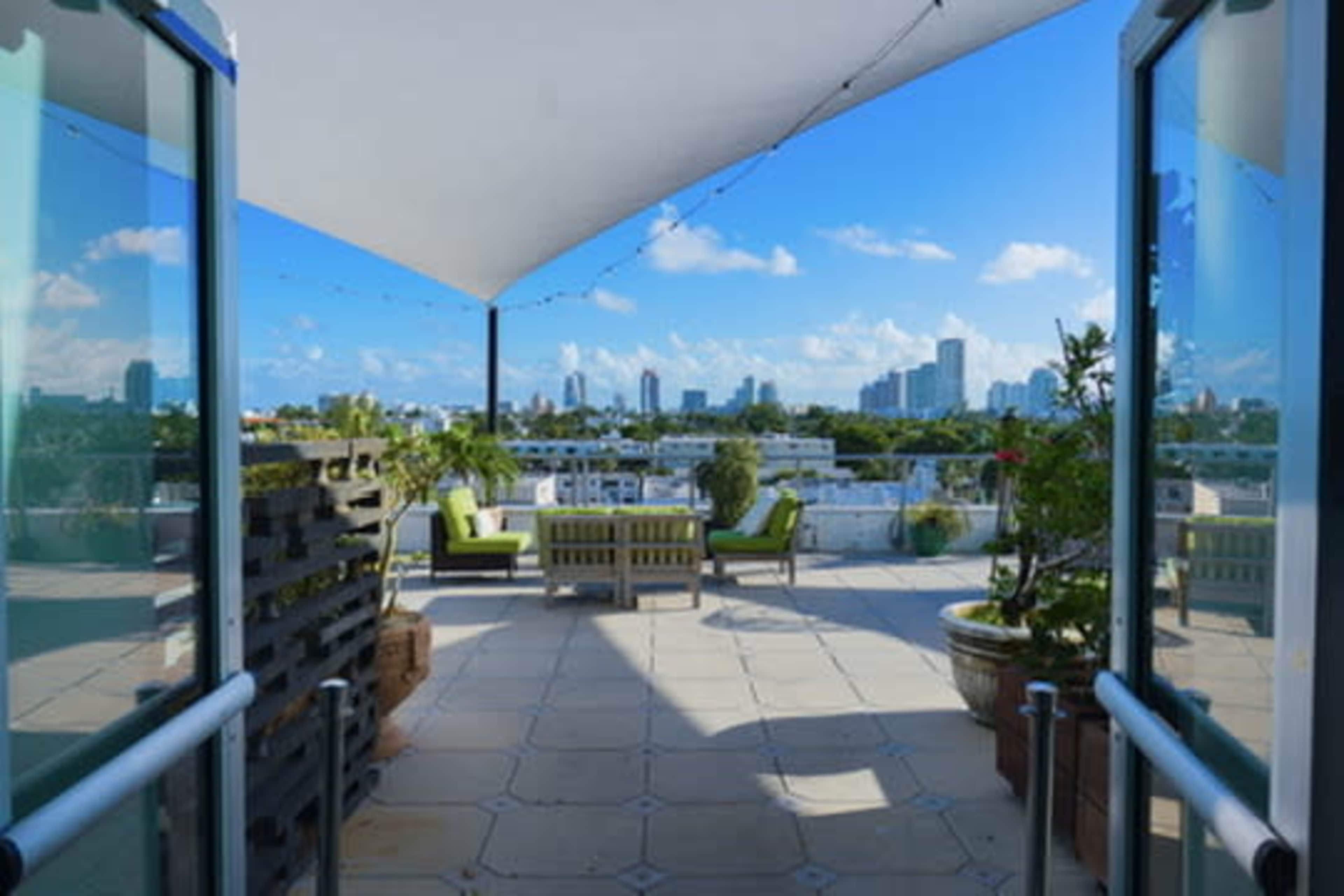 A rooftop terrace features lounge furniture and potted plants, offering a view of a city skyline under a clear blue sky.