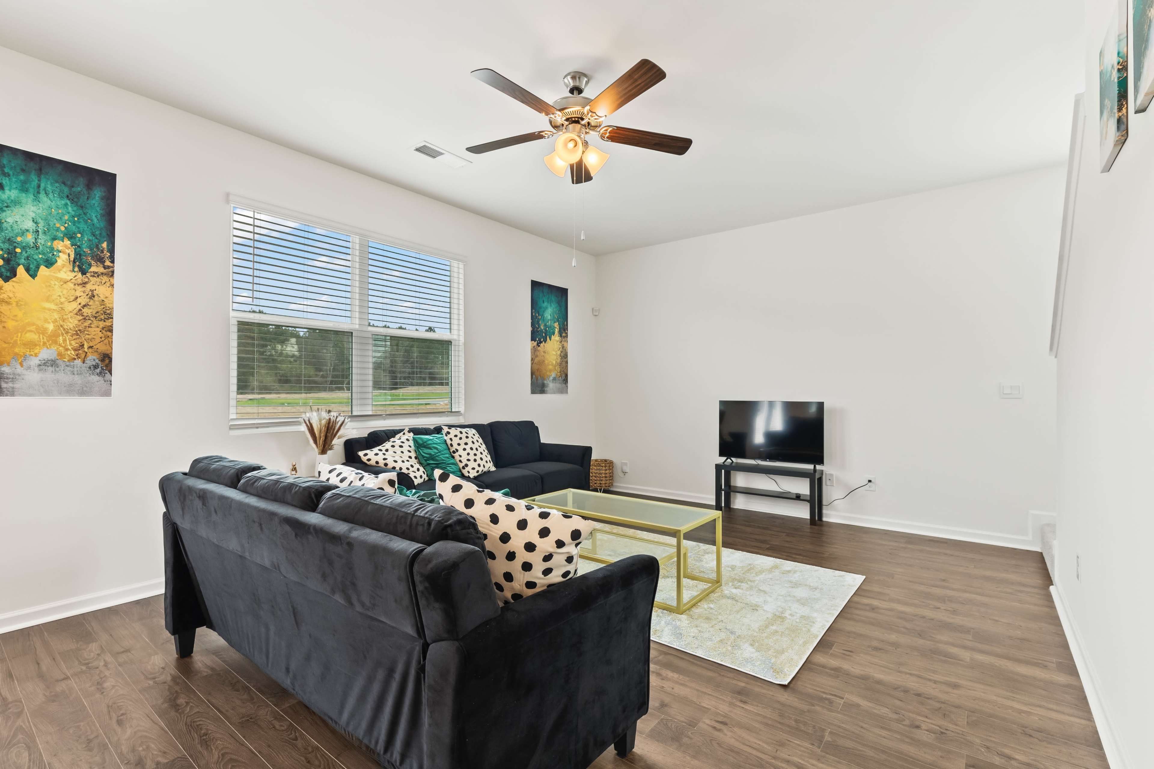 The living room features a black sofa, a glass coffee table, a television, and two artworks on the walls, with large windows allowing natural light.