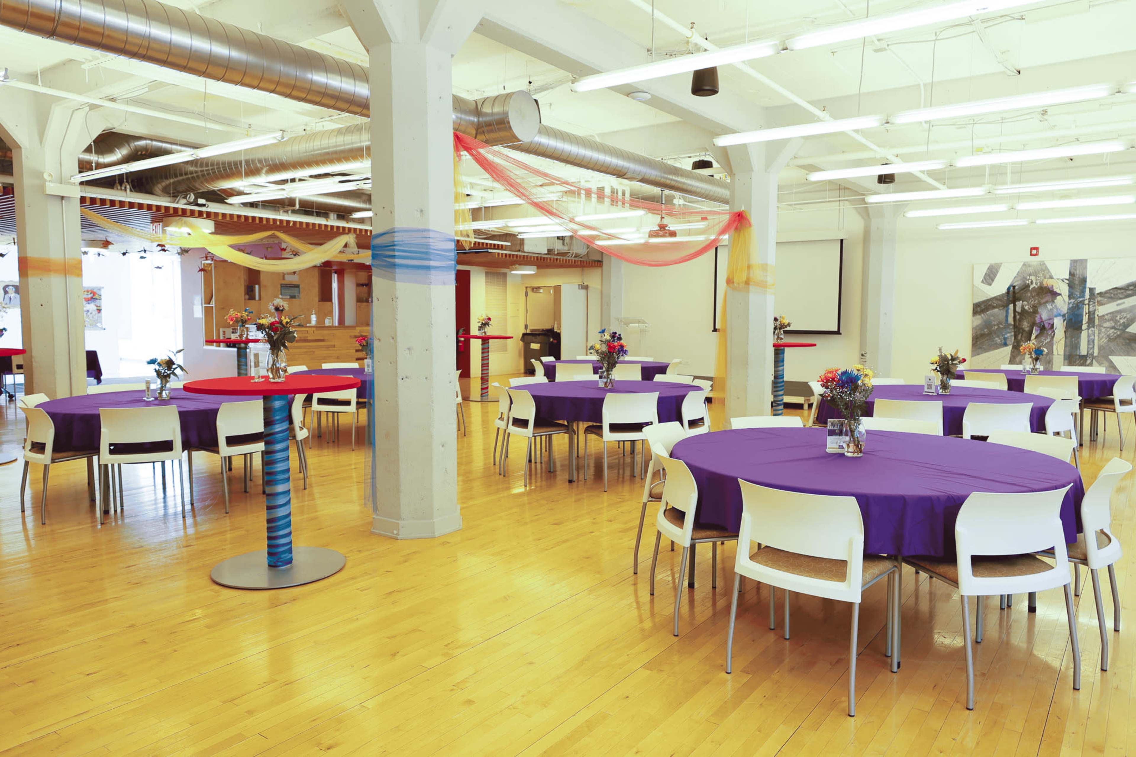 The image shows a spacious event venue with several round tables covered in purple tablecloths, each adorned with a small vase of flowers, set against a backdrop of modern interior design.