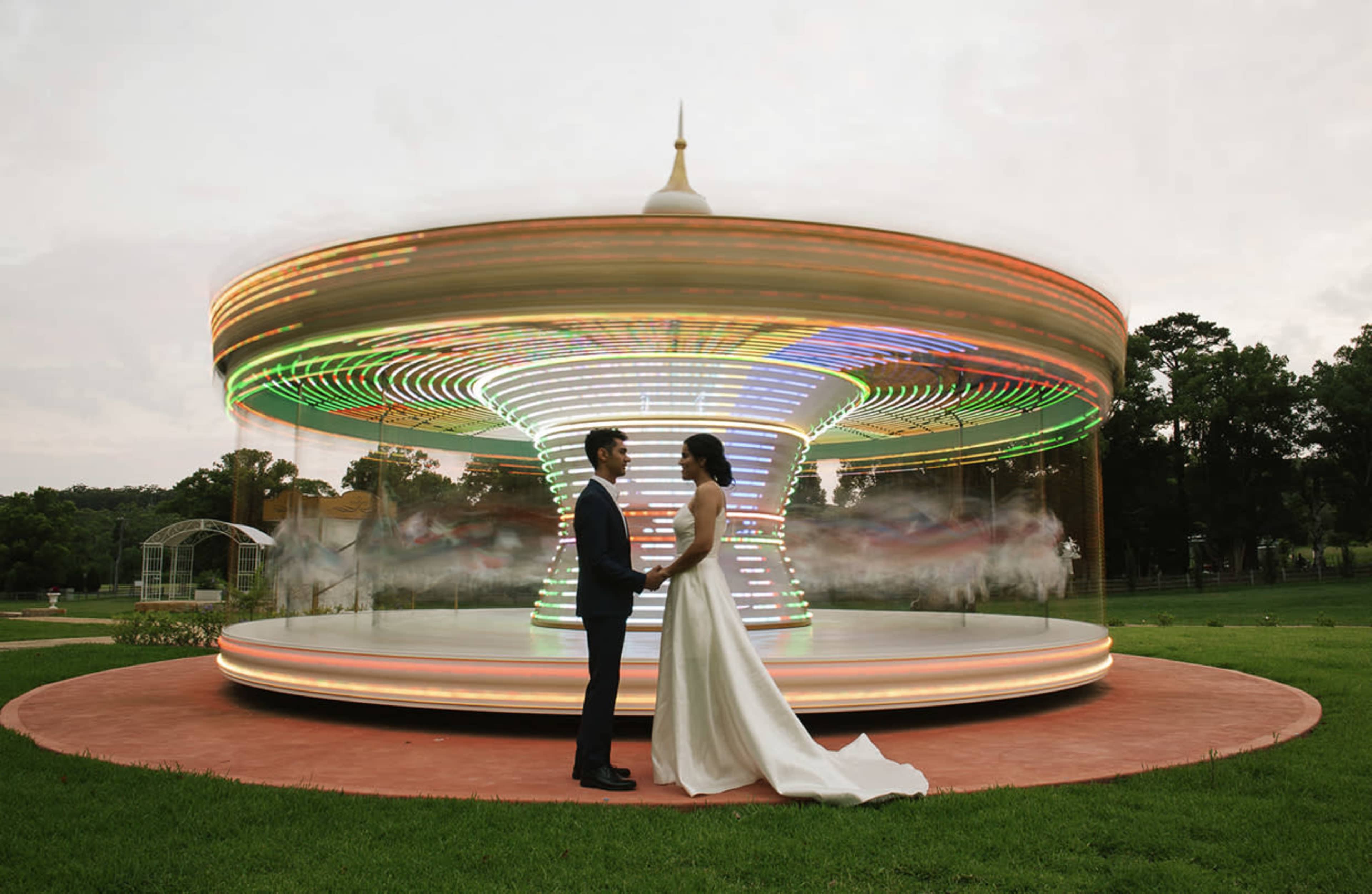 A couple stands facing each other in front of a brightly lit carousel in a green park.