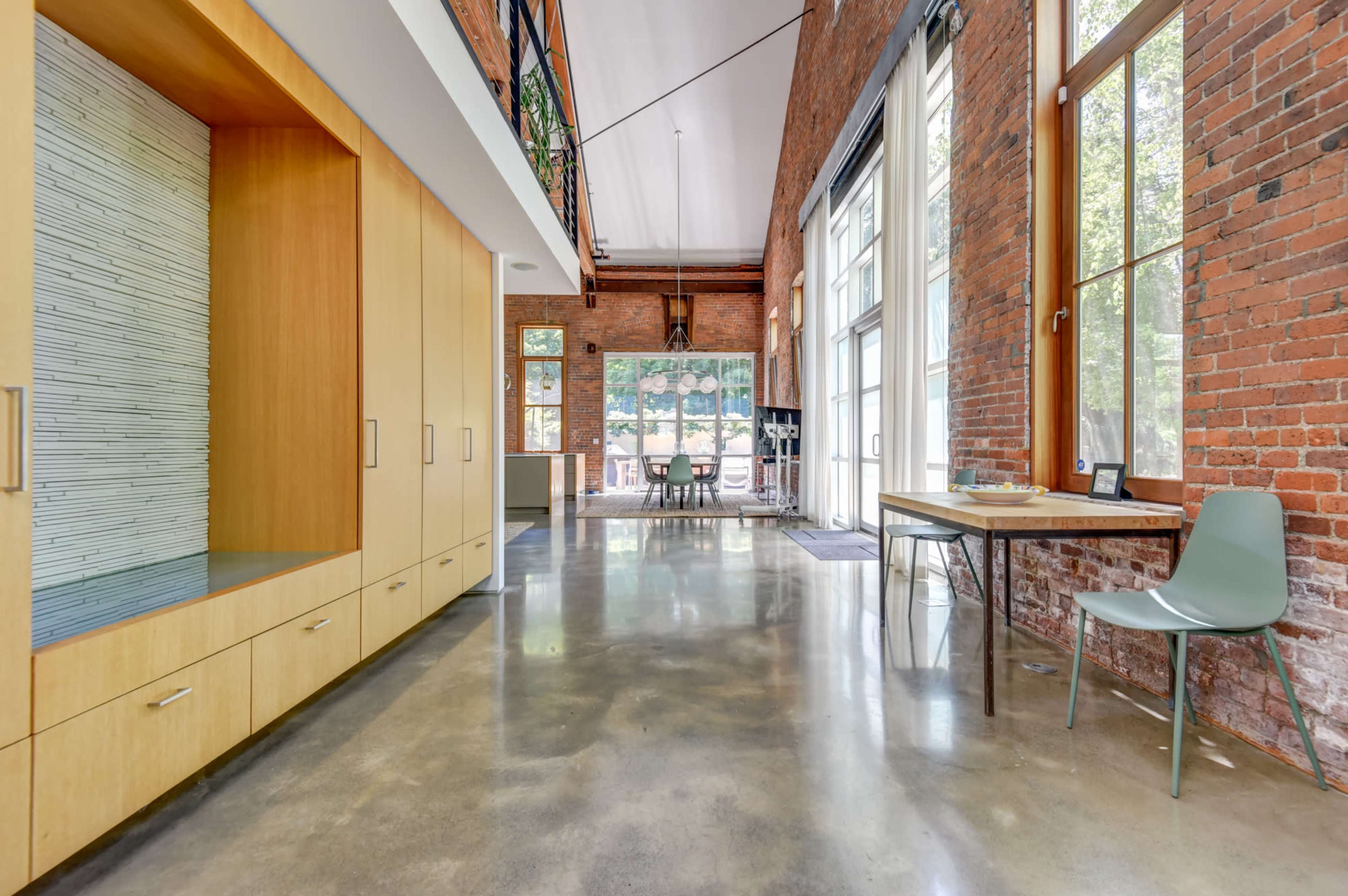 The image shows a modern, open-concept living space featuring exposed brick walls, polished concrete floors, and large windows that allow natural light to fill the room.