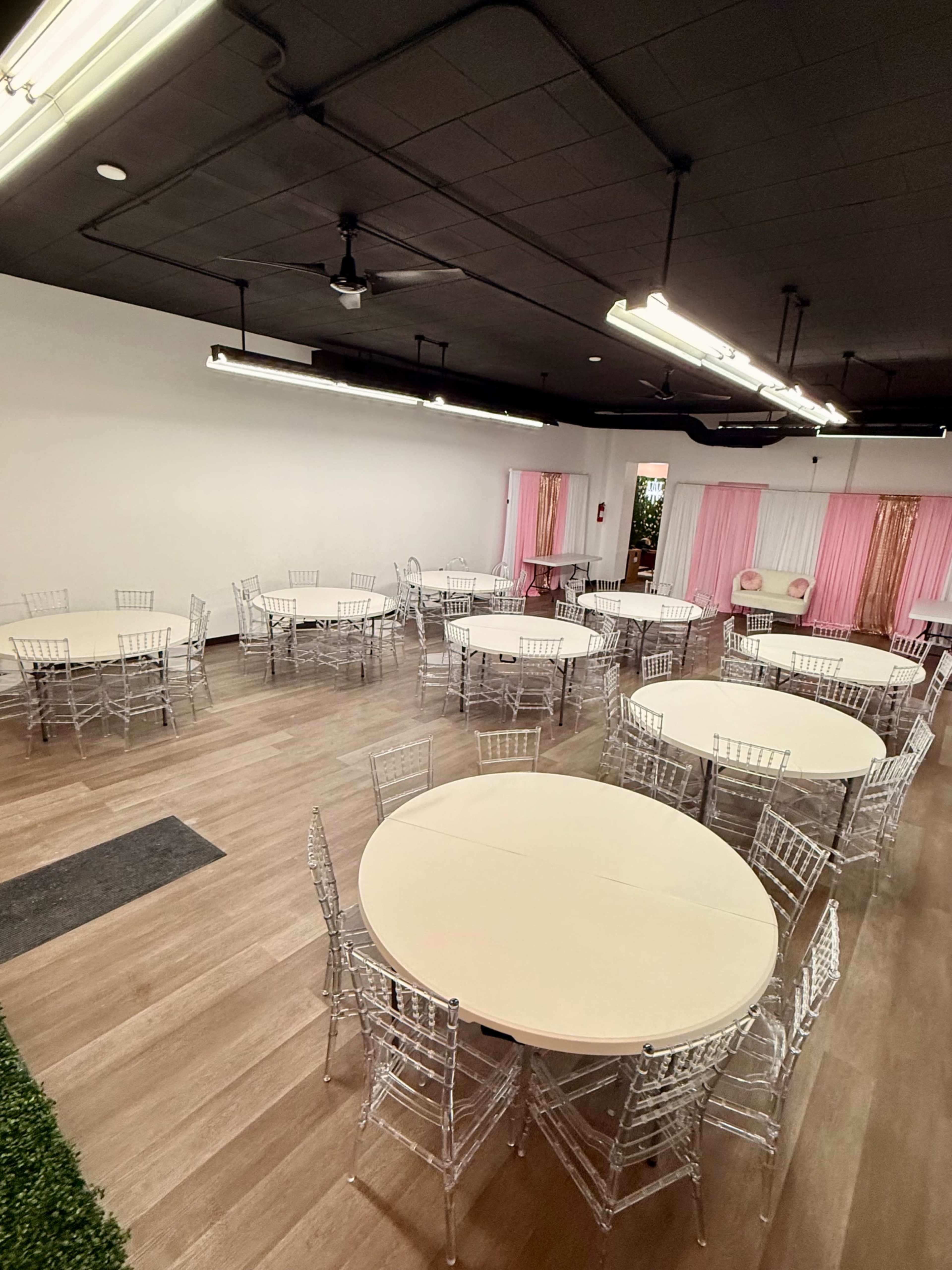 The image shows an event space with several round tables surrounded by clear chiavari chairs, arranged neatly on a hardwood floor.