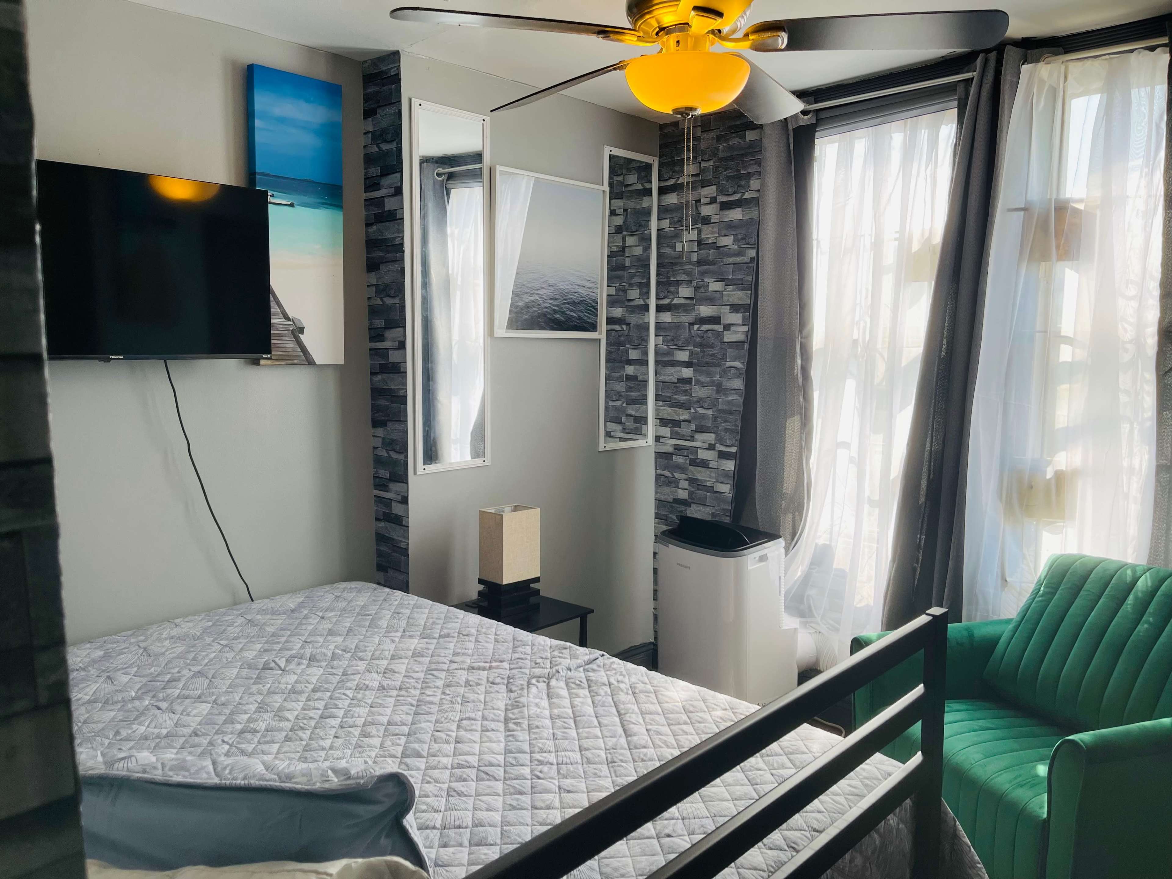 A modern bedroom features a bed, a green armchair, a television mounted on the wall, and large windows with drapes.