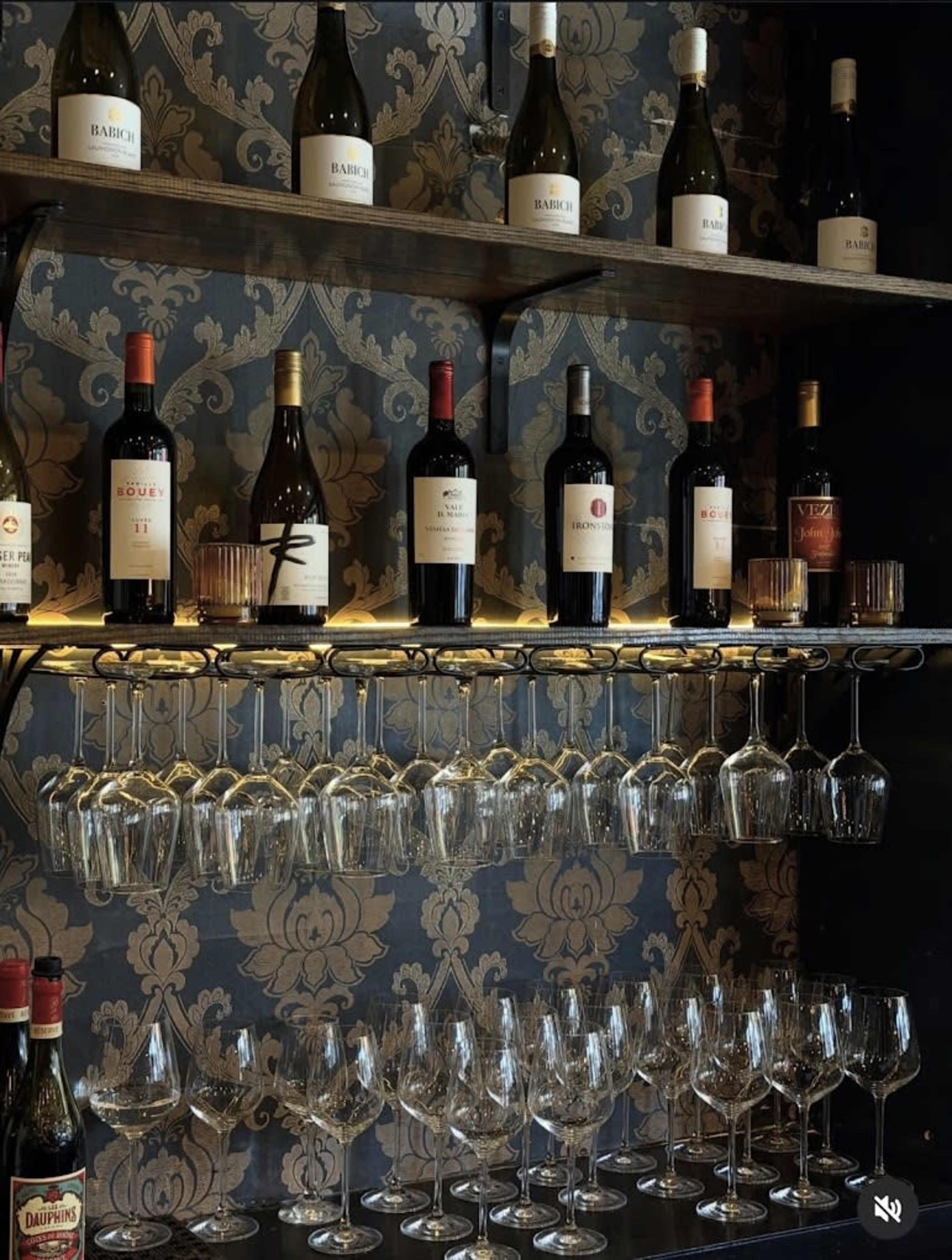 A wooden shelf displays various bottles of wine and champagne above a collection of wine glasses, set against a patterned wallpaper.