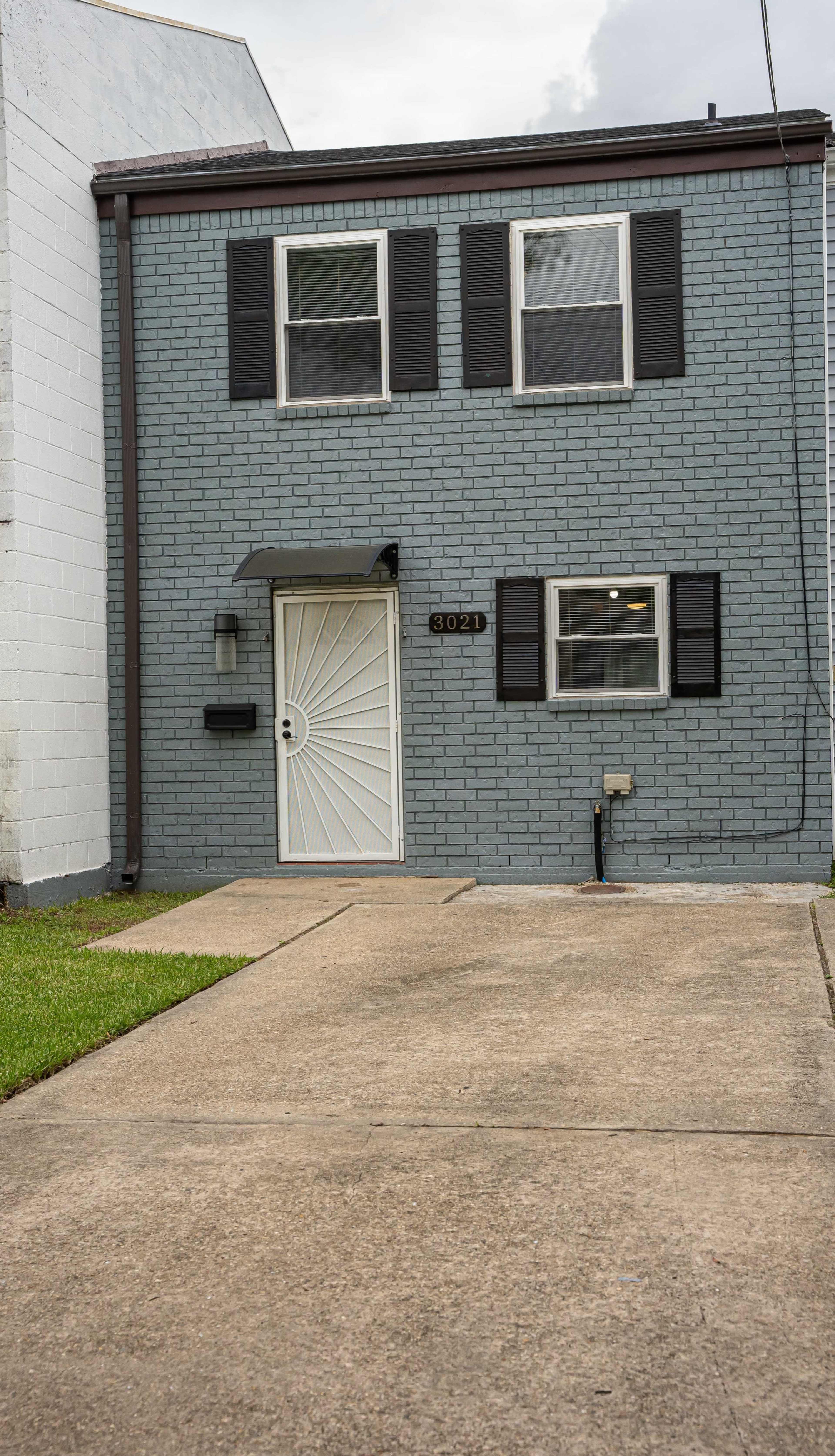 A residential building with a light gray brick exterior, featuring a front door with a decorative metal design and two windows framed by black shutters.