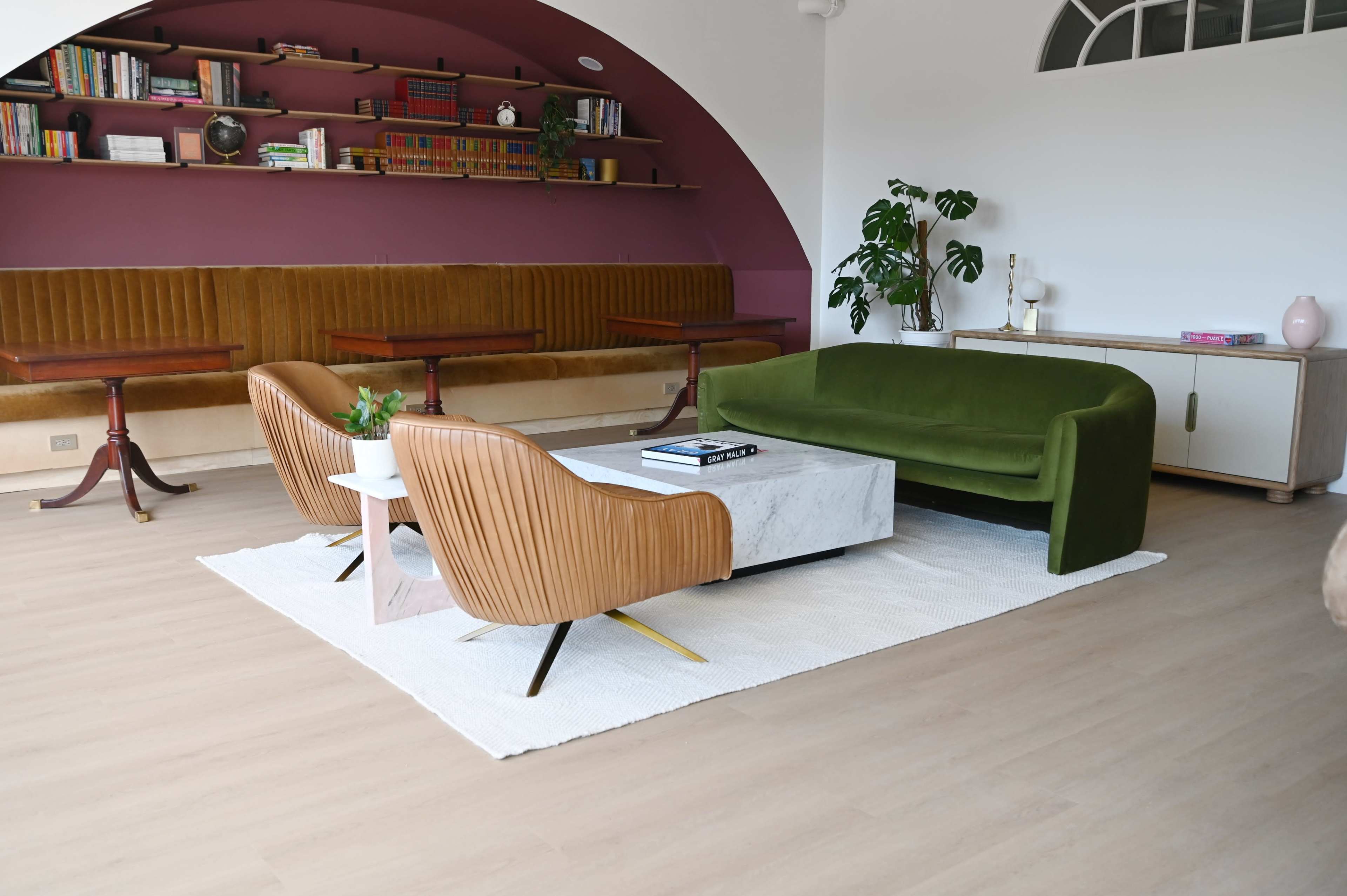 The image shows a modern living area featuring a green velvet sofa, two brown accent chairs, a marble coffee table, and a built-in bookshelf against a curved purple wall.