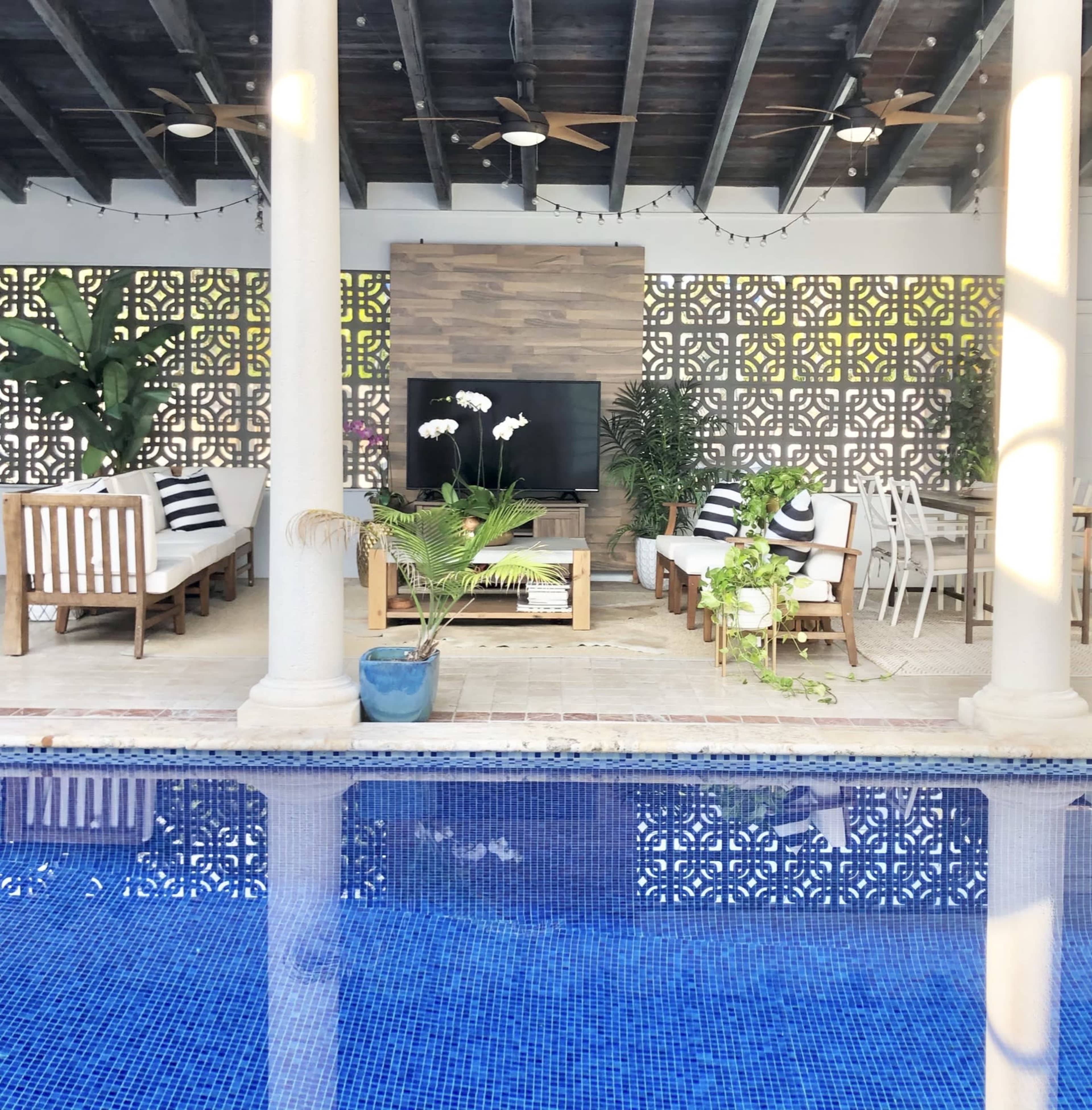 A patio area with seating, a television, and dining space overlooks a blue swimming pool.