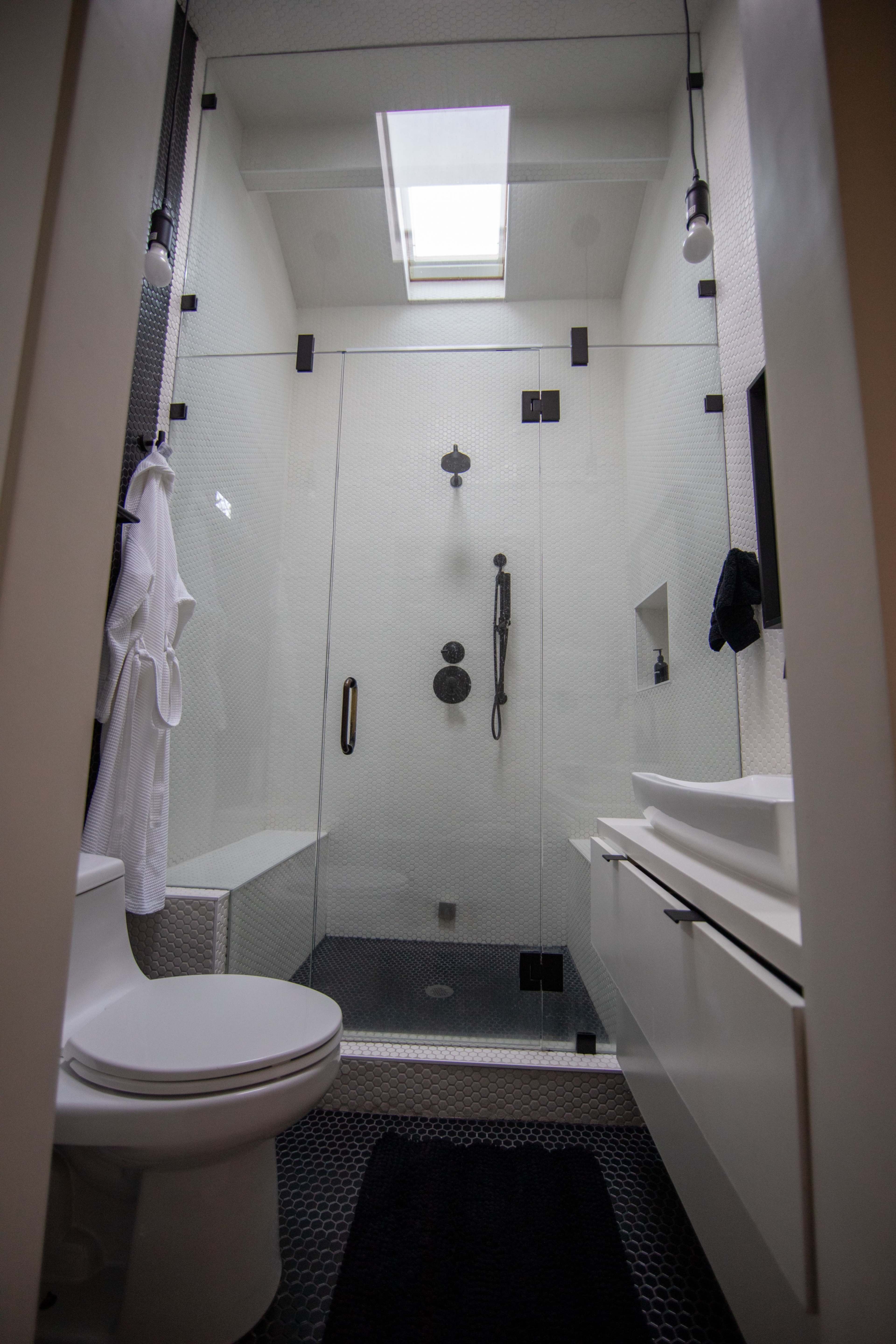 The image shows a modern bathroom with a glass shower enclosure, a toilet, a sink with a cabinet, and a skylight above.